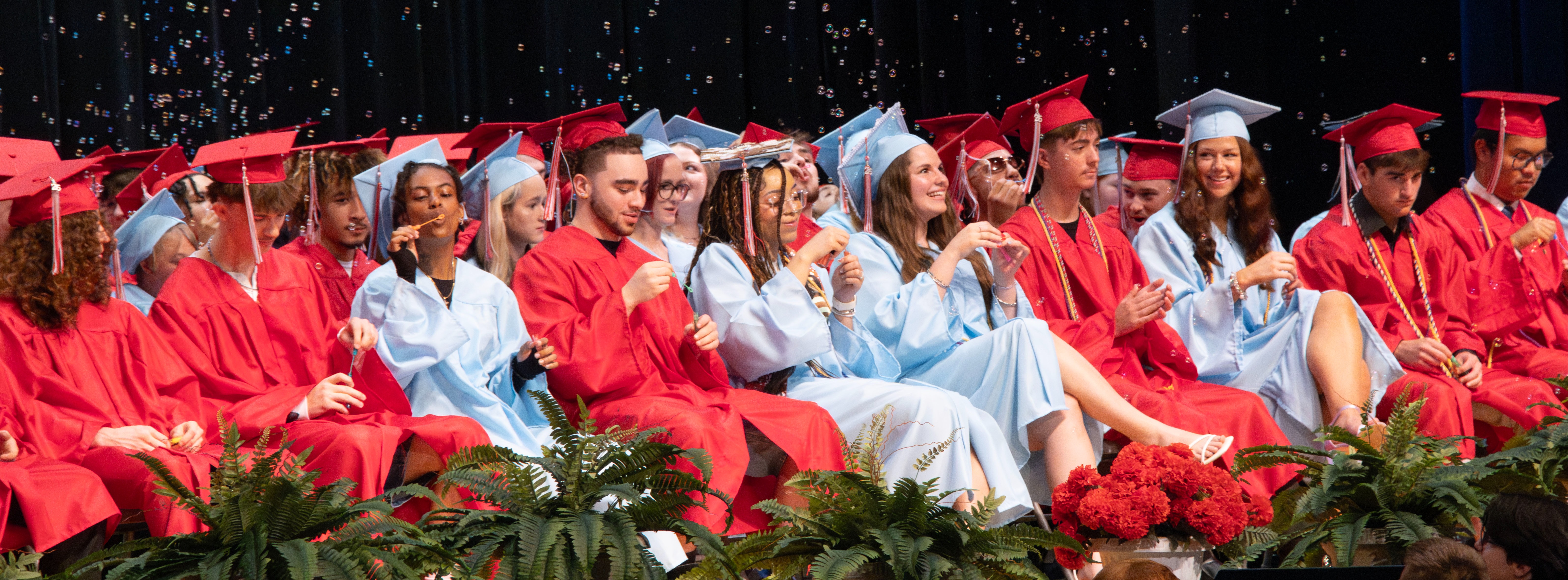High school graduates in red and Columbia blue caps and gowns sit together on stage as bubbles float through the air during their graduation ceremony.