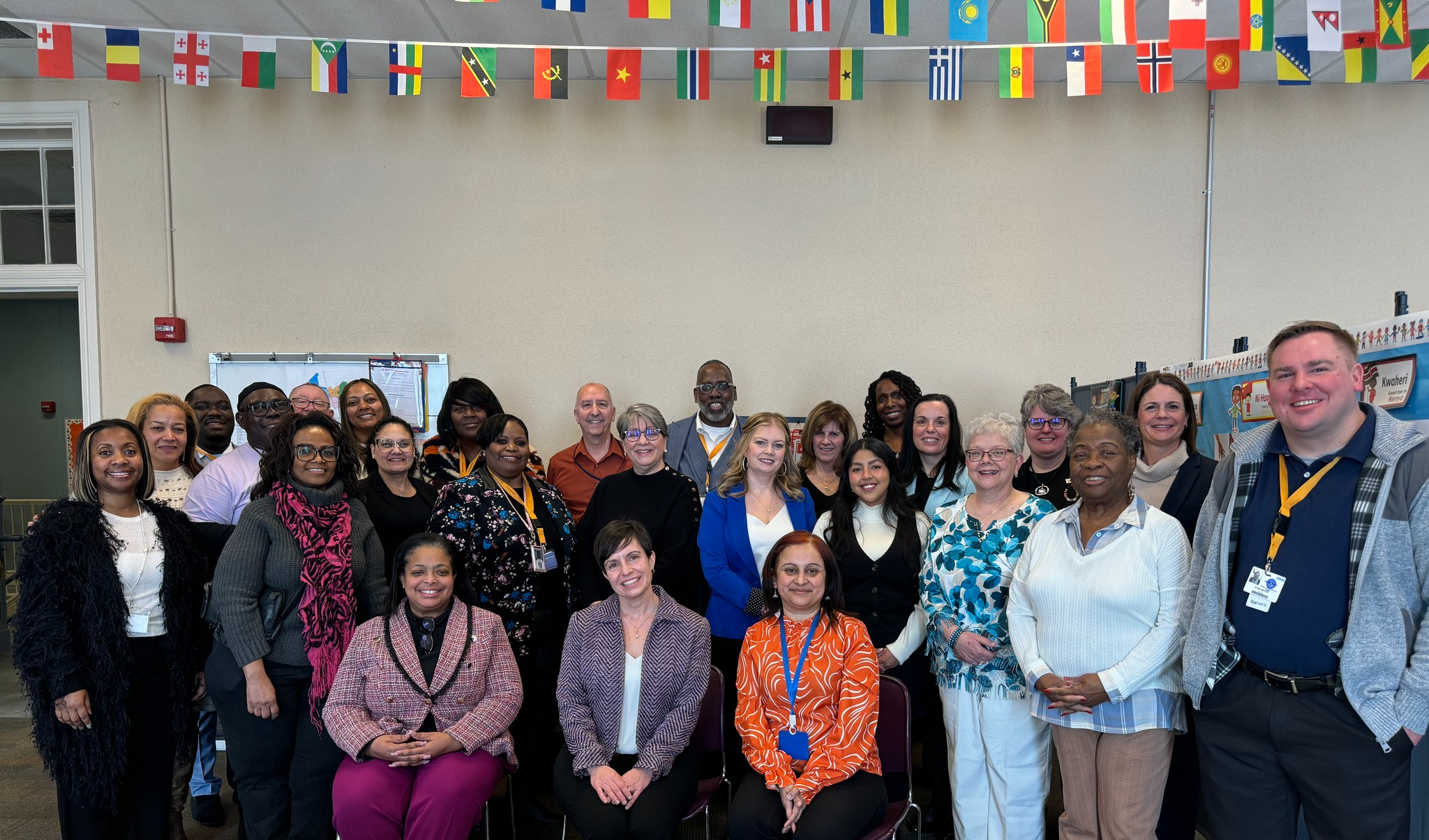 A diverse group of about thirty people posing for a group photo in a classroom setting with international flags hanging above. Some individuals are seated while others stand behind them.