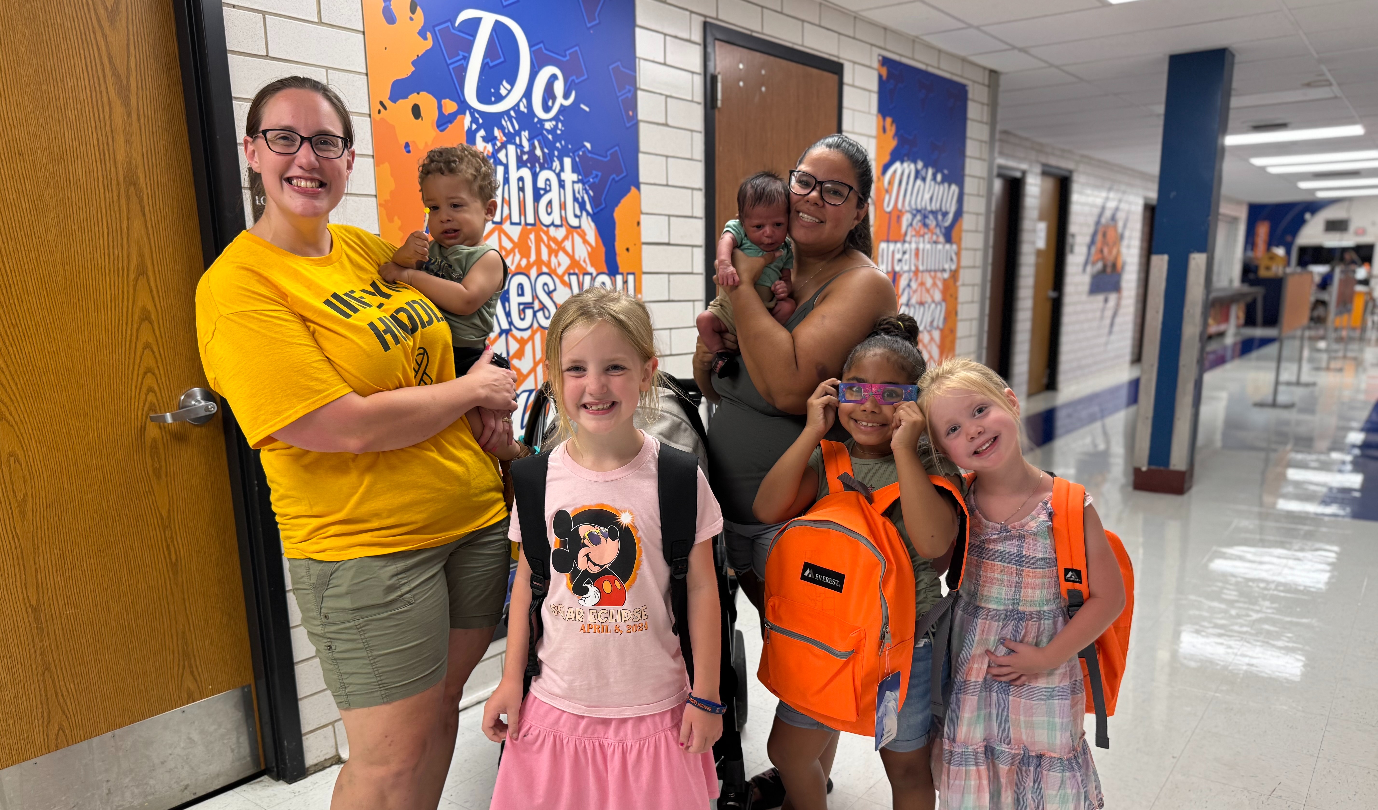 Two adults and five children are standing in a school cafeteria that has motivational posters in the background. The adults are holding two small children in their hands and the other three children are wearing backpacks.