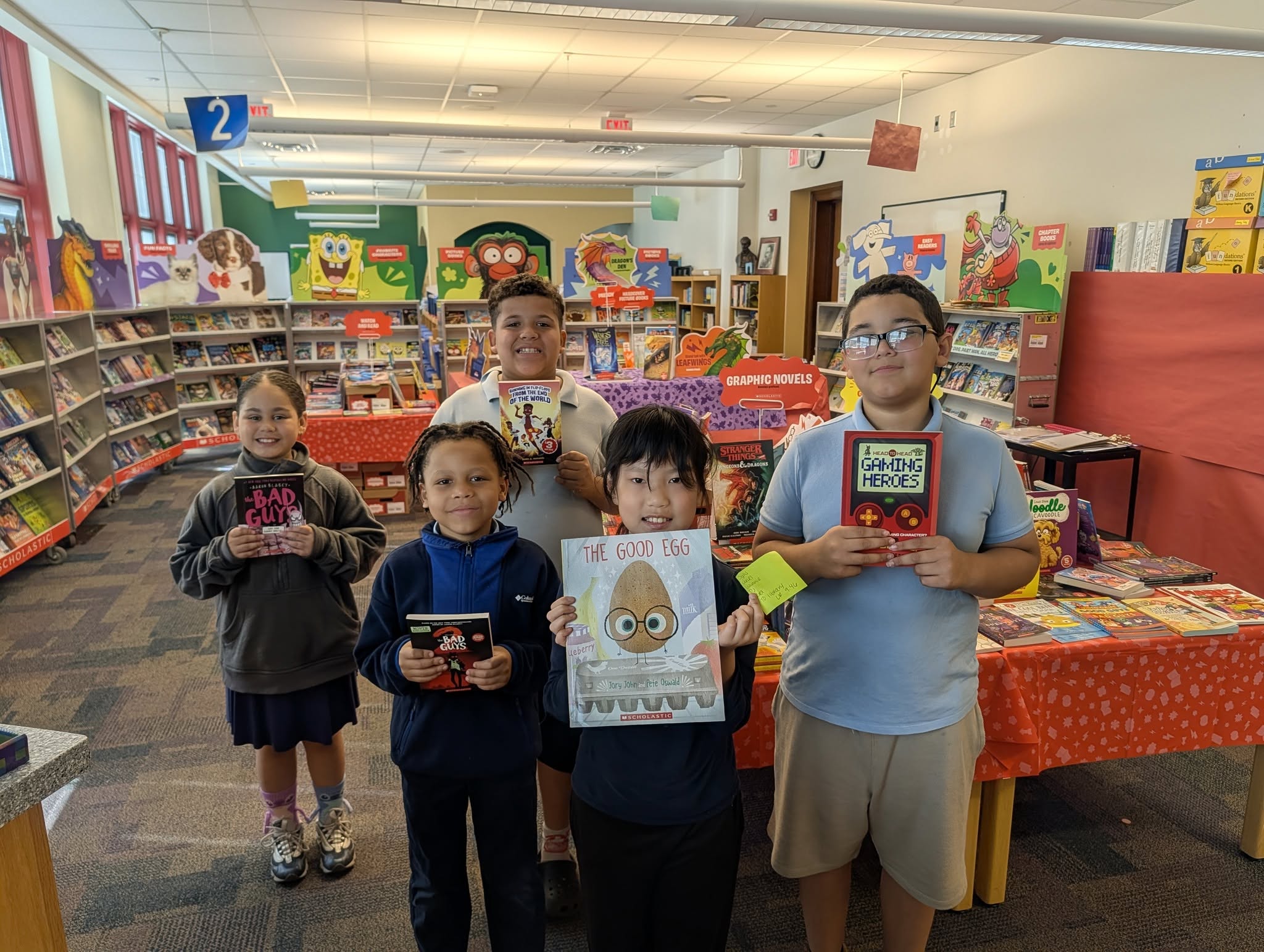 Five students are standing together at a school book fair, smiling and holding up books.