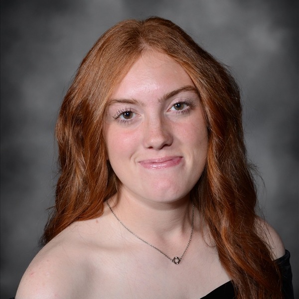 Senior Portrait of Crystal Williams. She is wearing a black wrap slightly off the shoulders. Her red hair is curled and hanging on her left side. She is wearing a delicate gold chain necklace with a small pendant. The portrait is just a headshot, form the chest up.