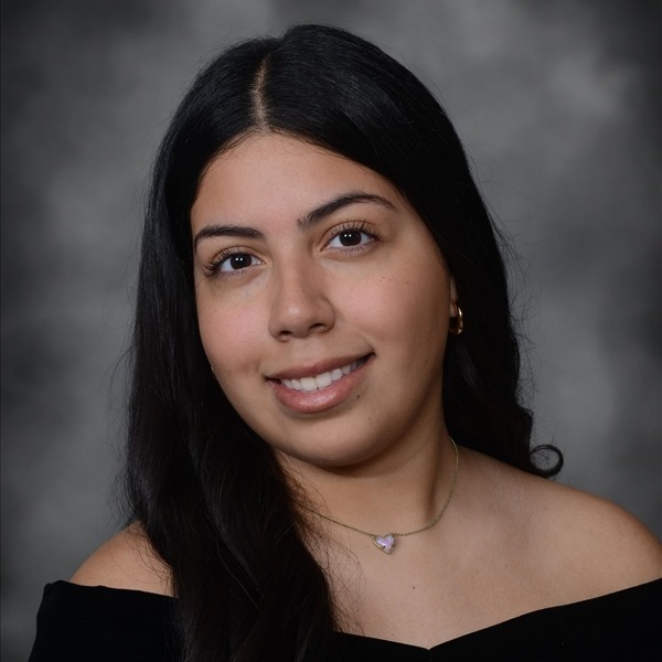 Senior Portrait of Rosa Villegas-Ramirez. She is wearing a black wrap slightly off the shoulders. Her dark brown hair is curled and hanging on her right side. She is wearing a delicate gold chain necklace with a small white heart pendant. She is also wearing small gold hoop earrings.The portrait is just a headshot, form the chest up.