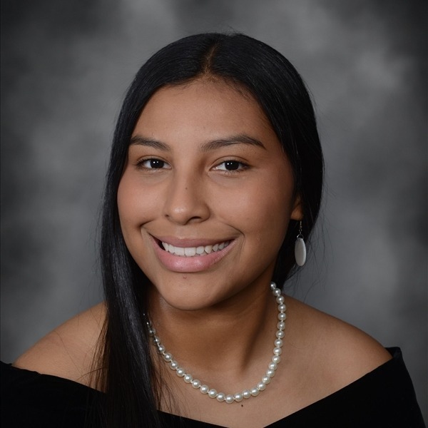 Senior Portrait of Danika Bullcoming. She is wearing a black wrap slightly off the shoulders. Her dark hair is straight and hanging on her right side. She has a pearl necklace on and white earrings. The portrait is just a headshot, form the chest up.
