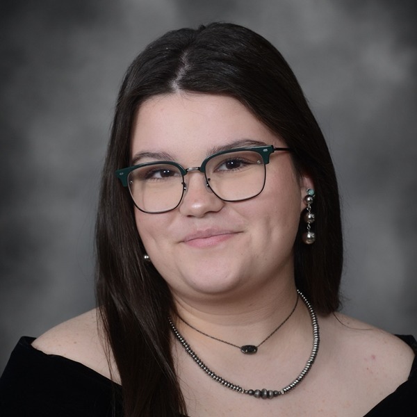 Senior Portrait of Kira Harris. She is wearing a black wrap slightly off the shoulders. Her dark hair is straight and hanging on her right side. She is wearing a double strand necklace and hanging earrings with three hanging gold balls. She is wearing a dark green and black framed pair of glasses.The portrait is just a headshot, form the chest up.