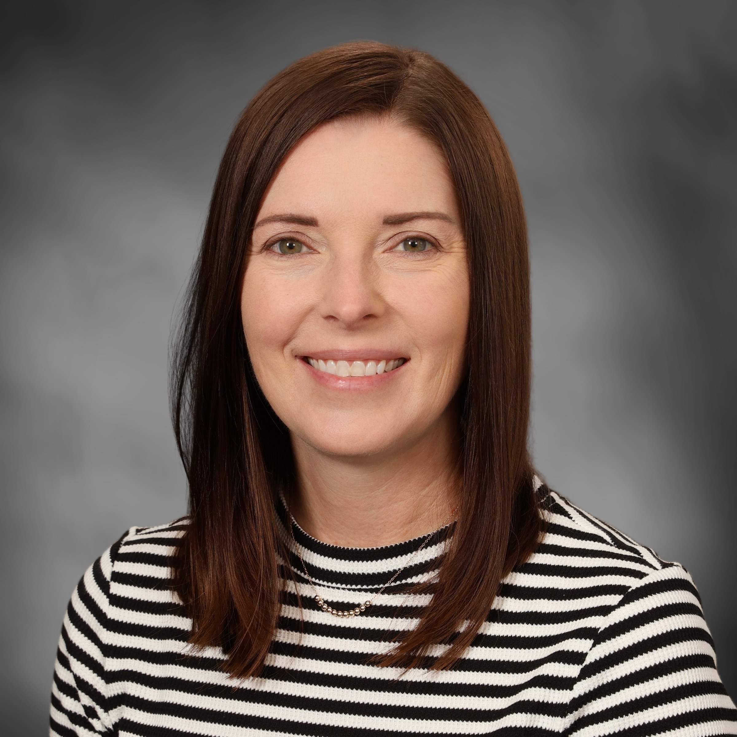 A woman with brown hair smiles at the camera. She wears a black and white striped shirt and a necklace.