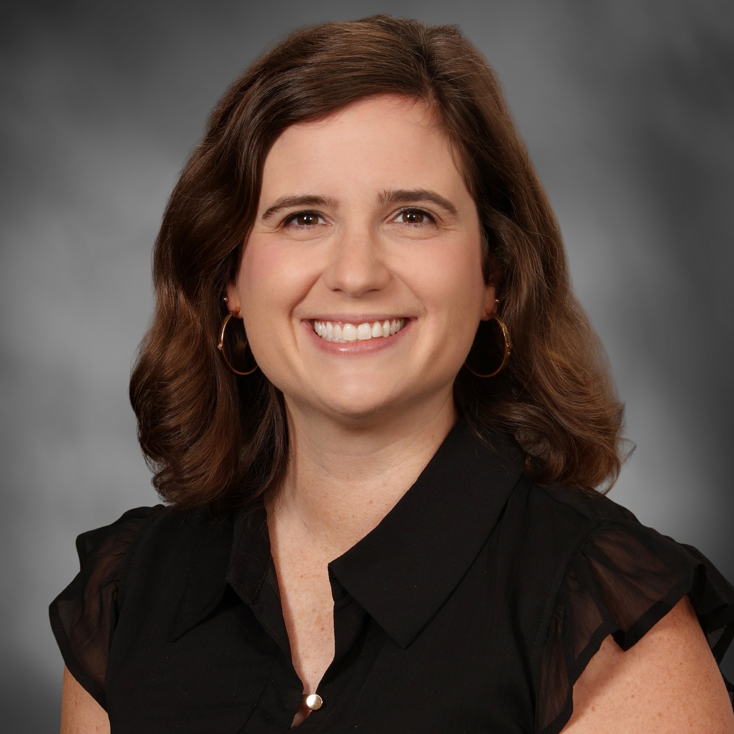 A woman with brown hair, smiling, wearing a black top with short sleeves. The background is gray.