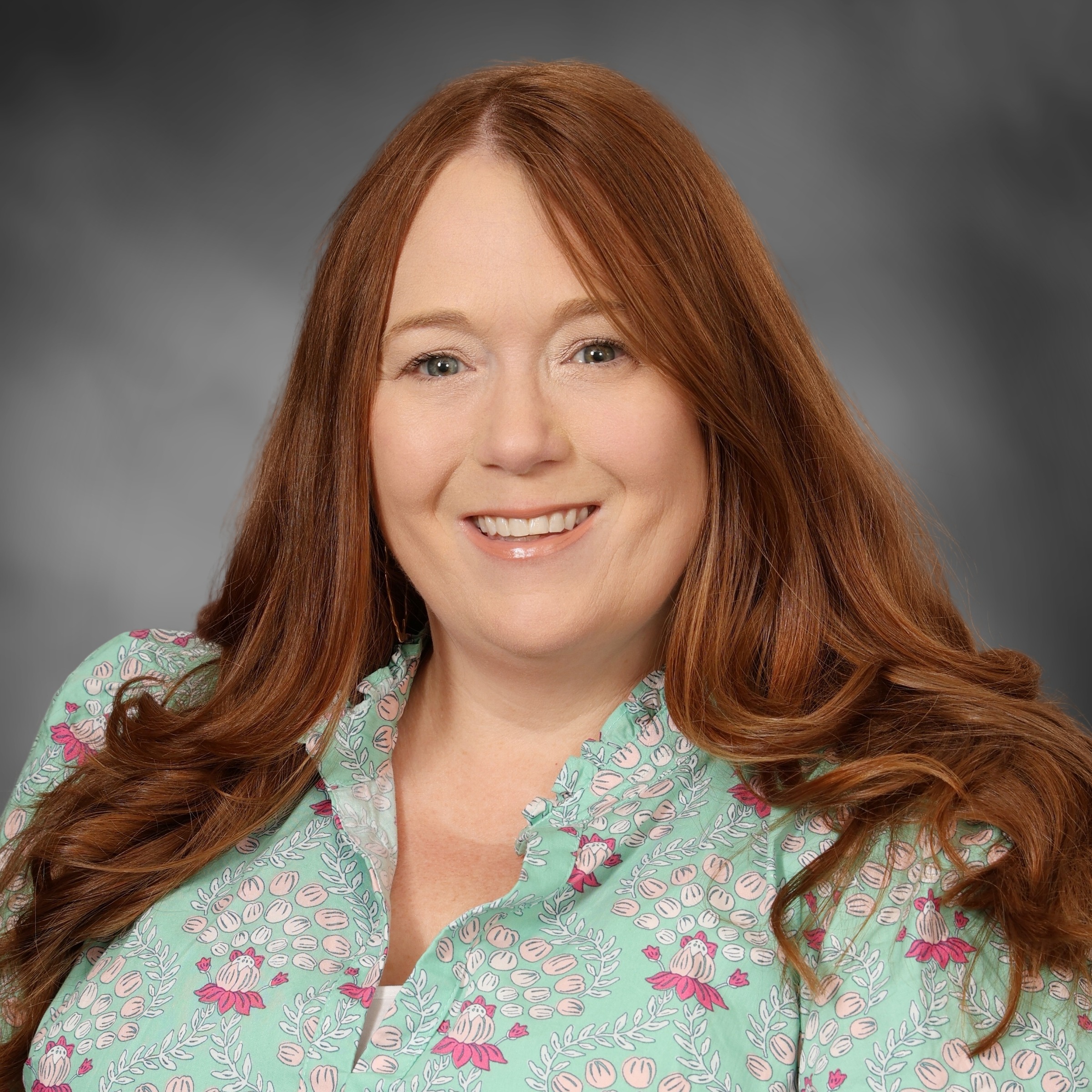 A woman with long curly brown hair and a green floral blouse, smiling and looking directly at the camera.