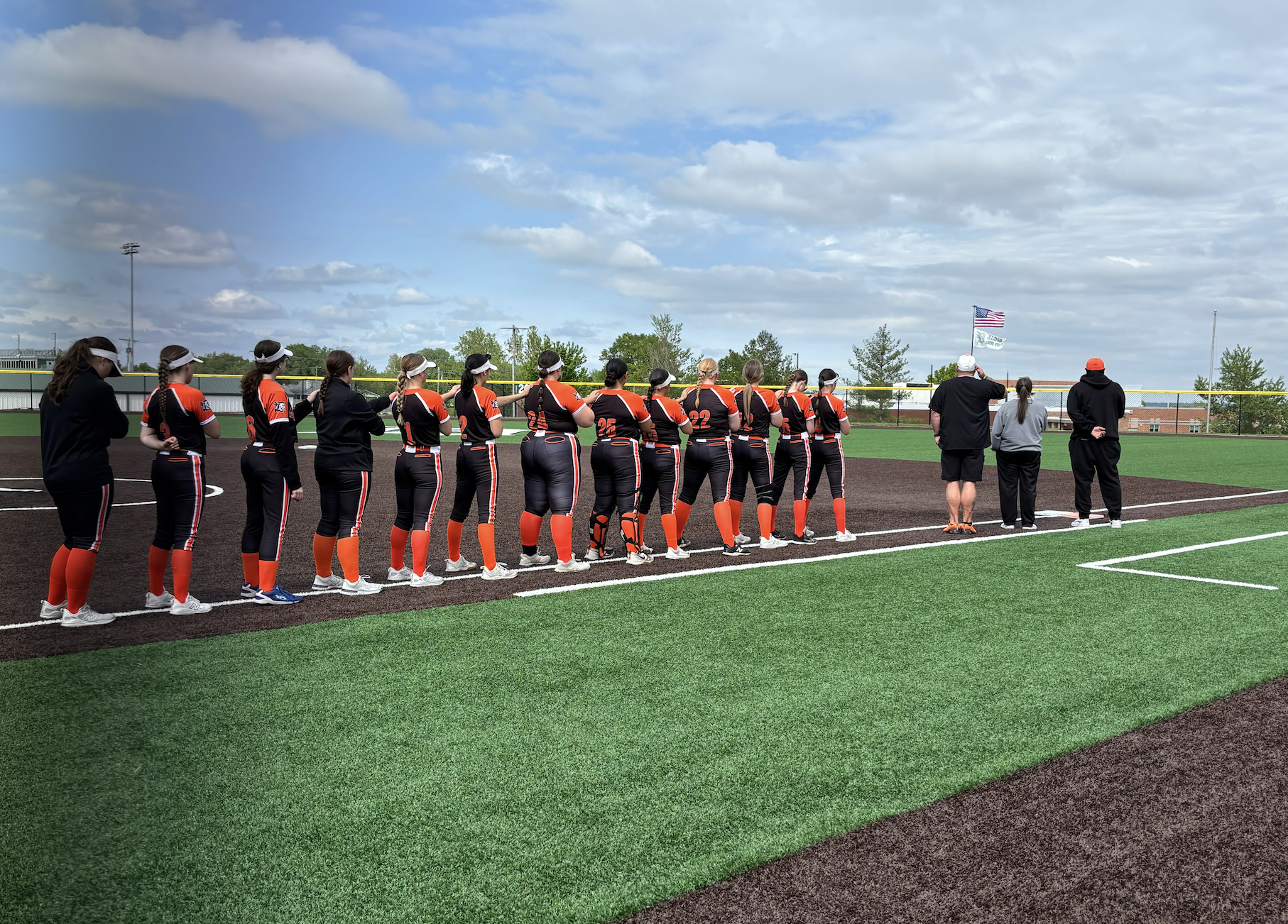 A team in black and orange uniforms stands on a field. Spectators stand nearby. Cloudy sky in the background.