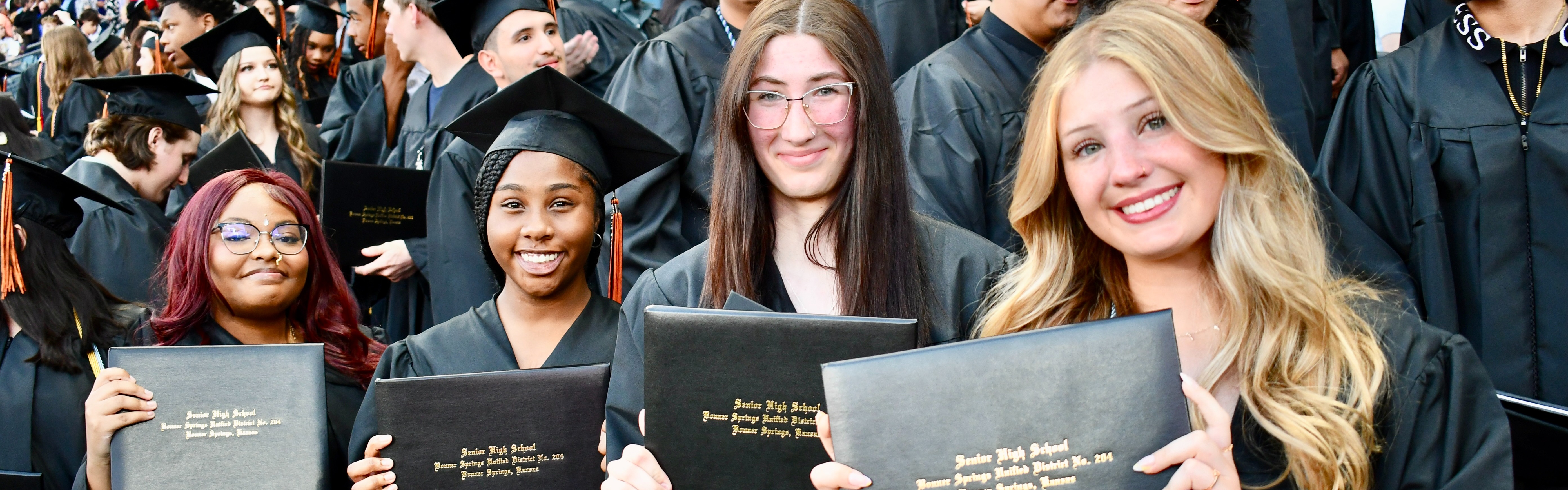 Five women in caps and gowns pose for a photo, holding their diplomas. Behind them, a crowd of graduates.