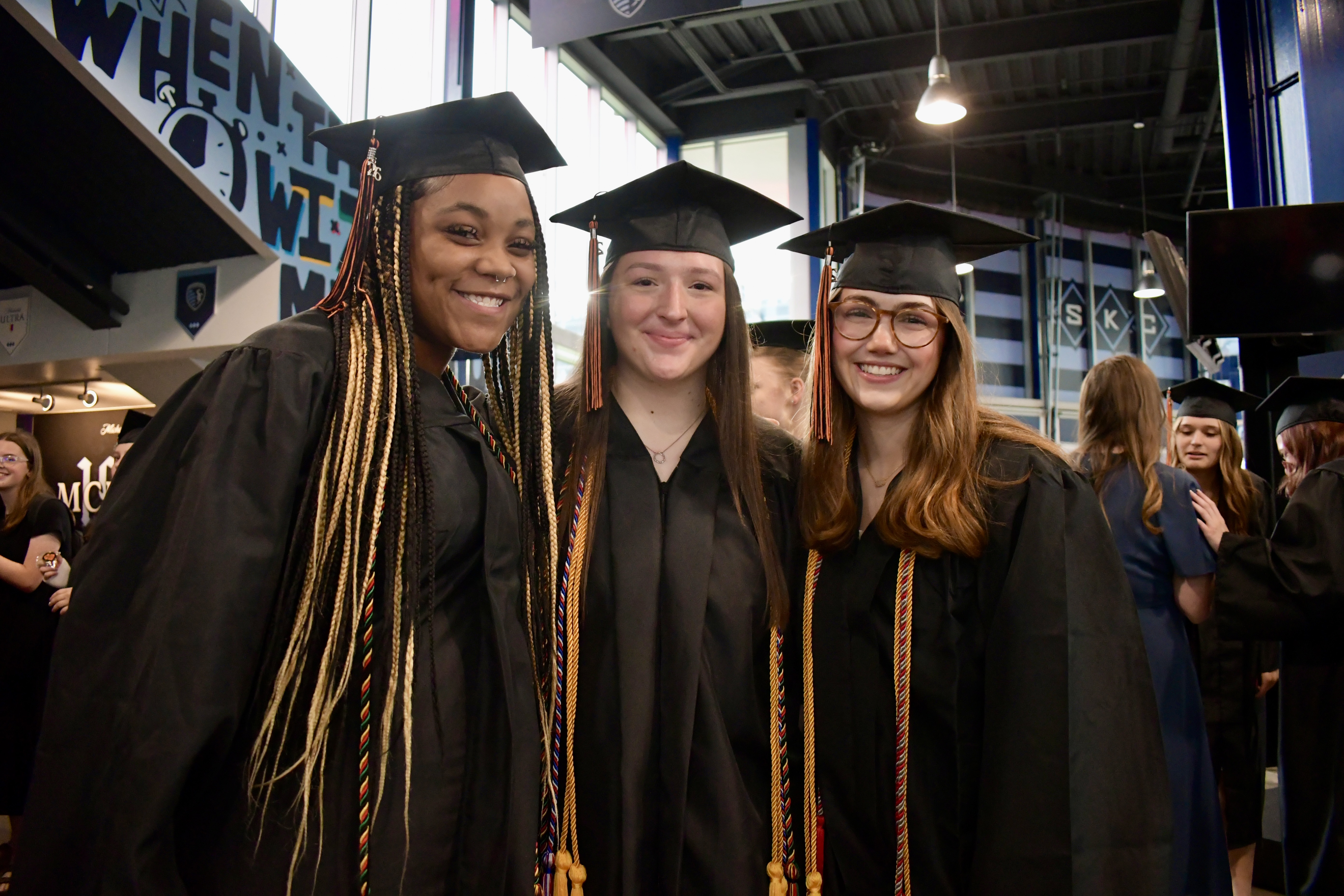 Three women in black graduation gowns and hats pose for a picture at an indoor event.