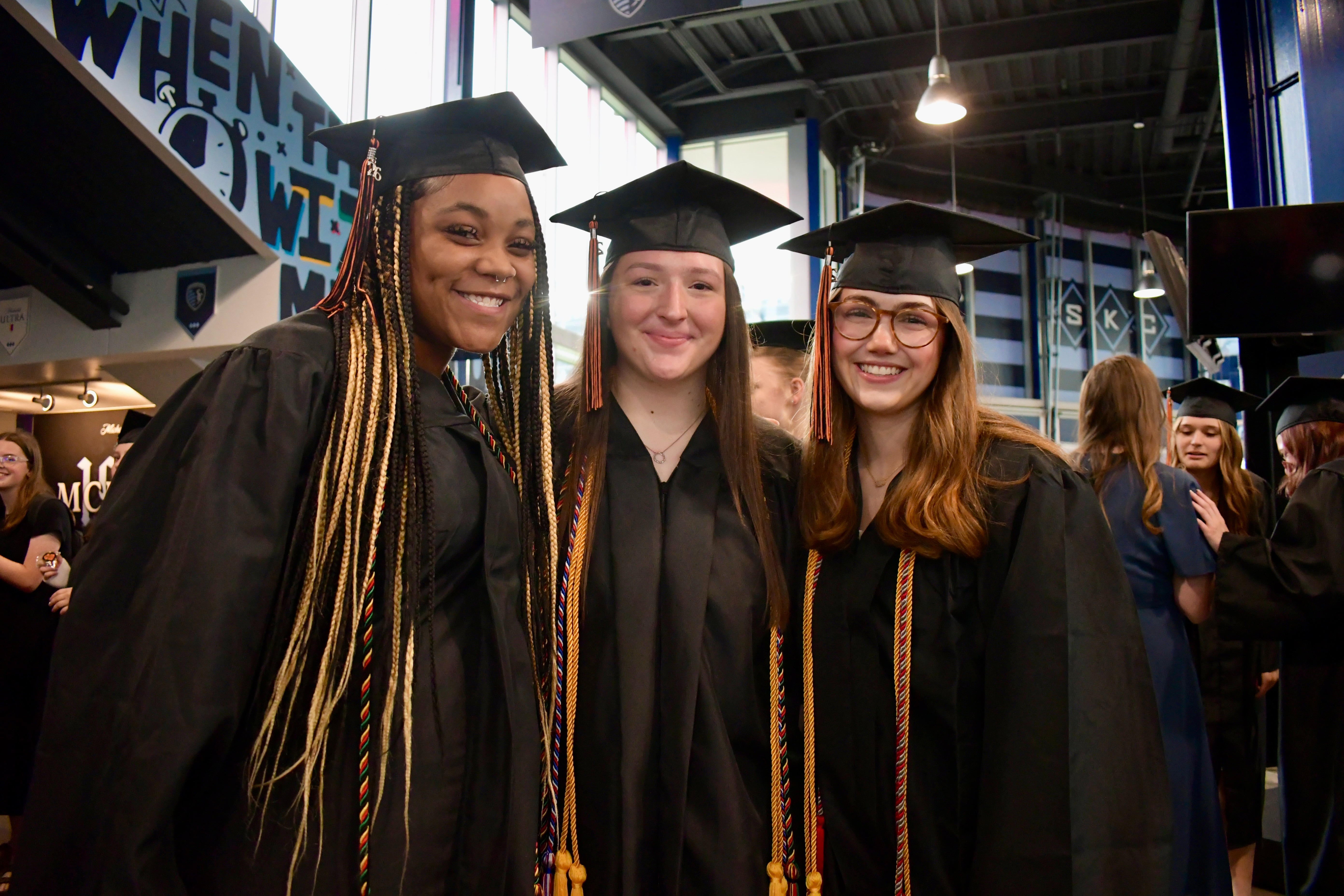 Three women in graduation gowns smile at camera, standing together in a crowded indoor hall.