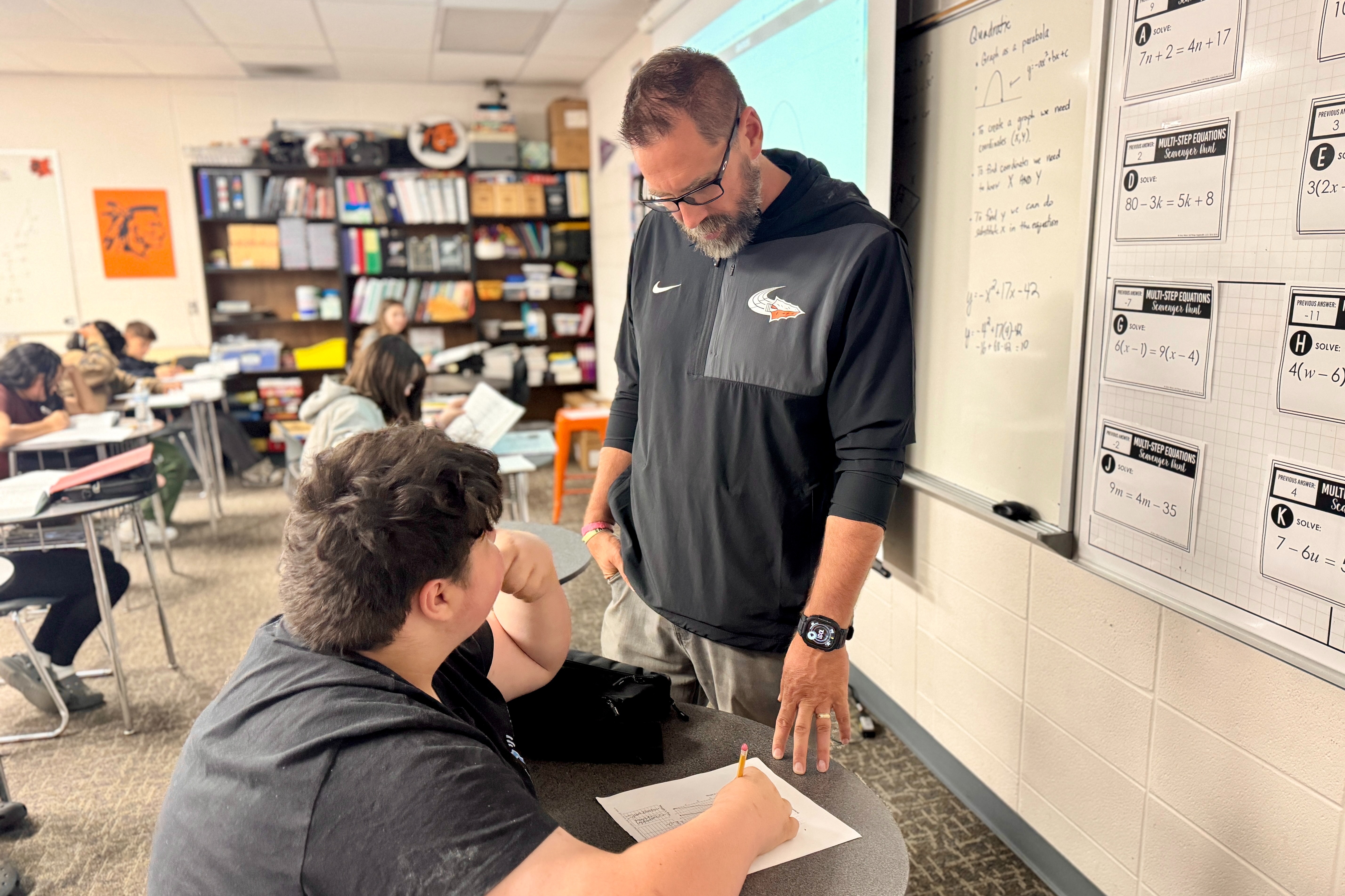 A man in a black jacket stands behind a student writing on paper at a desk in a classroom.