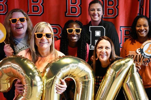 Several women standing behind gold foil balloons spelling 2024, against a red backdrop with letters B and R.