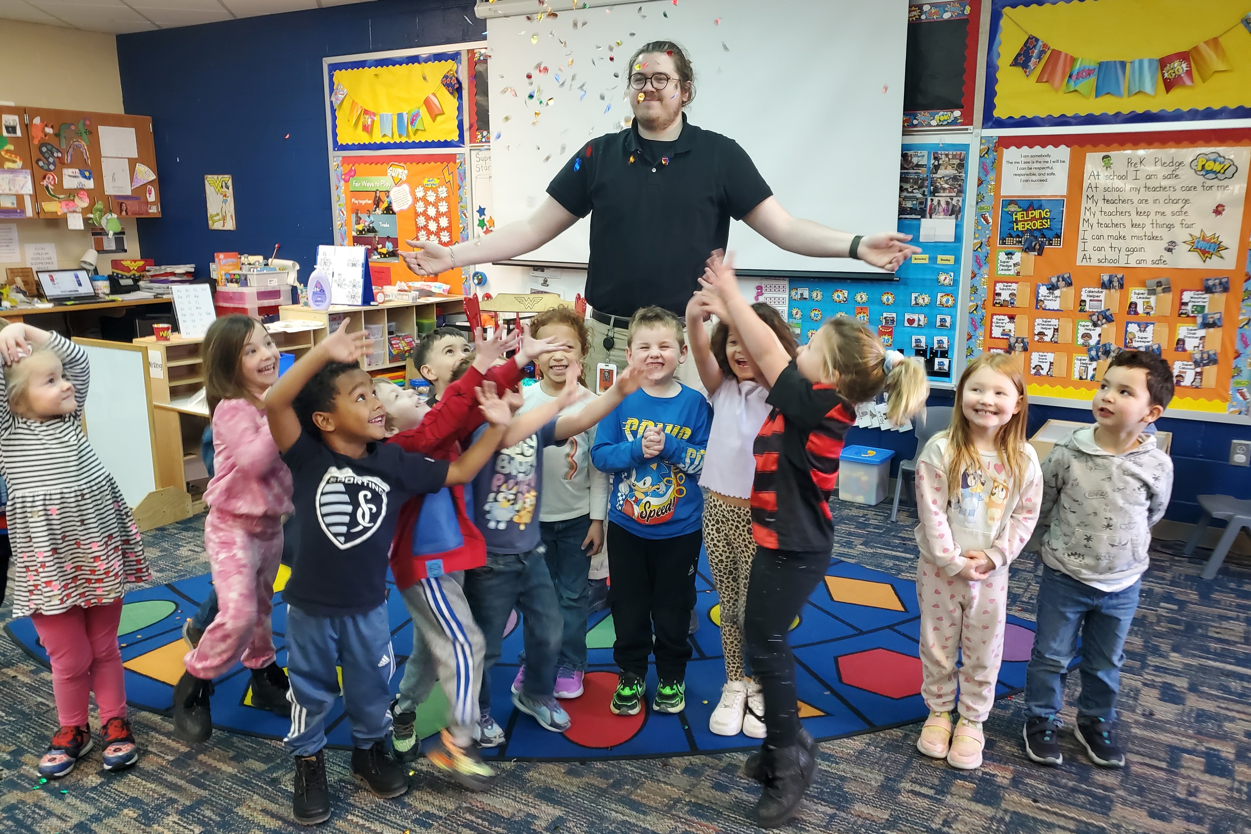 A classroom with children in colorful clothes dancing in a circle with a teacher.