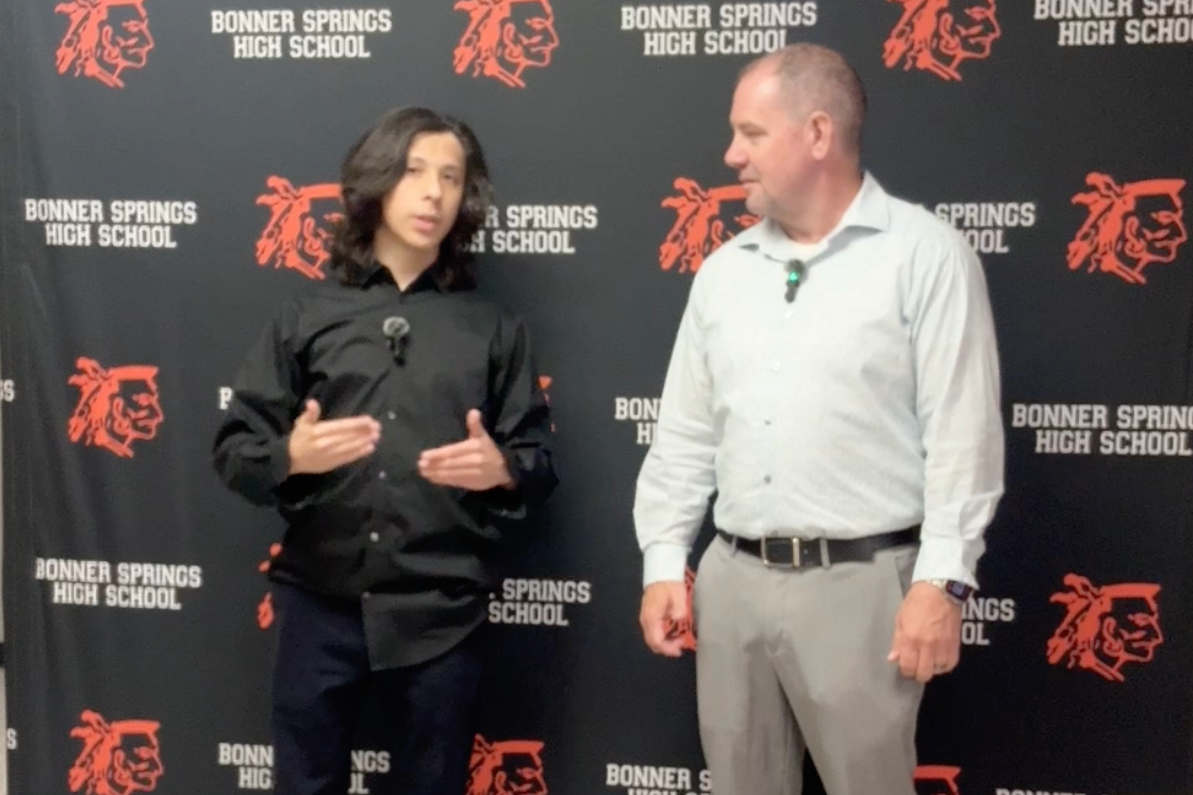 Two people stand in front of a backdrop with the Bonner Springs High School logo. One speaks into a microphone.
