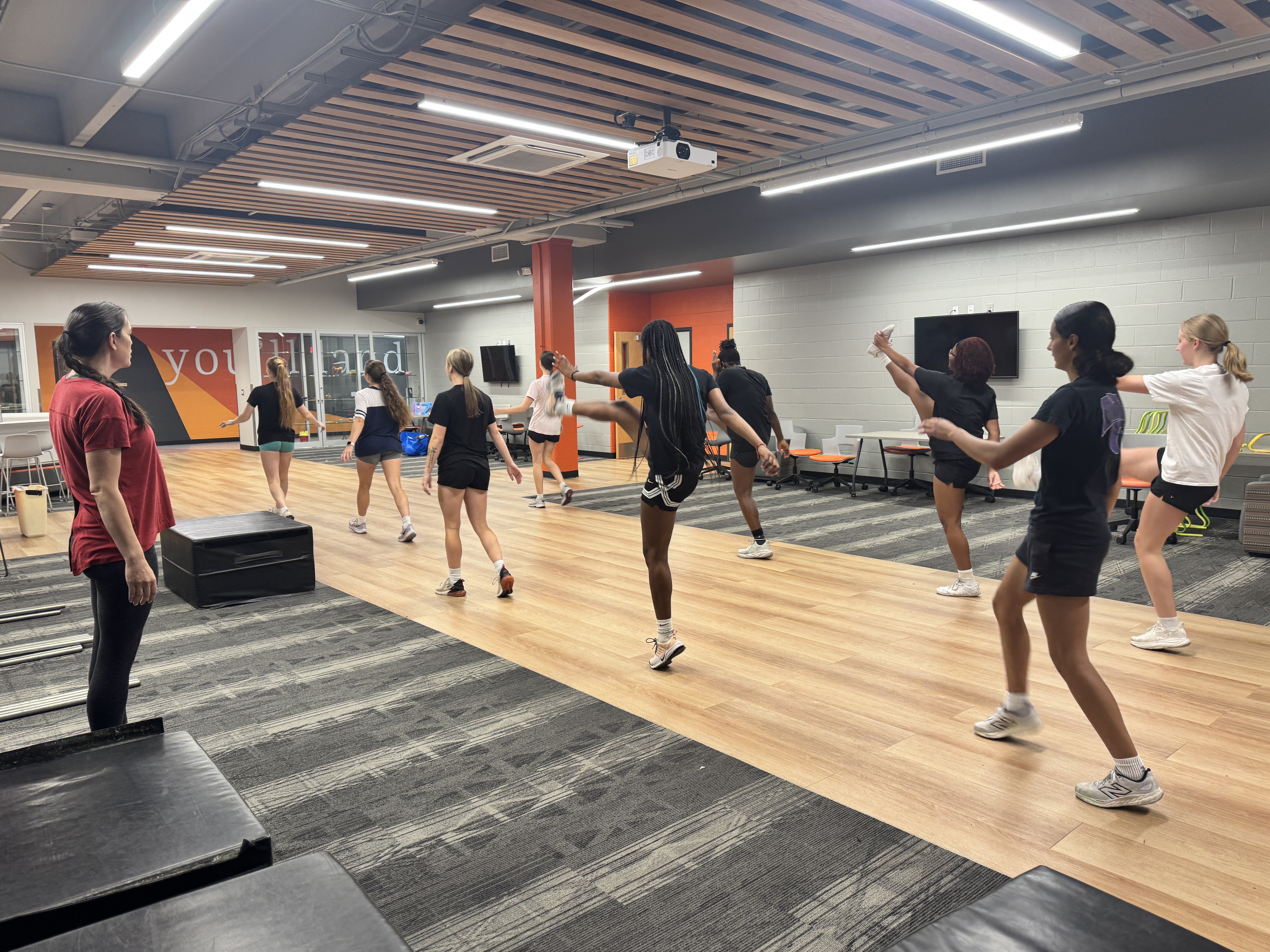 Several people in a gym practice dance moves on a wood floor; some are standing on exercise mats.