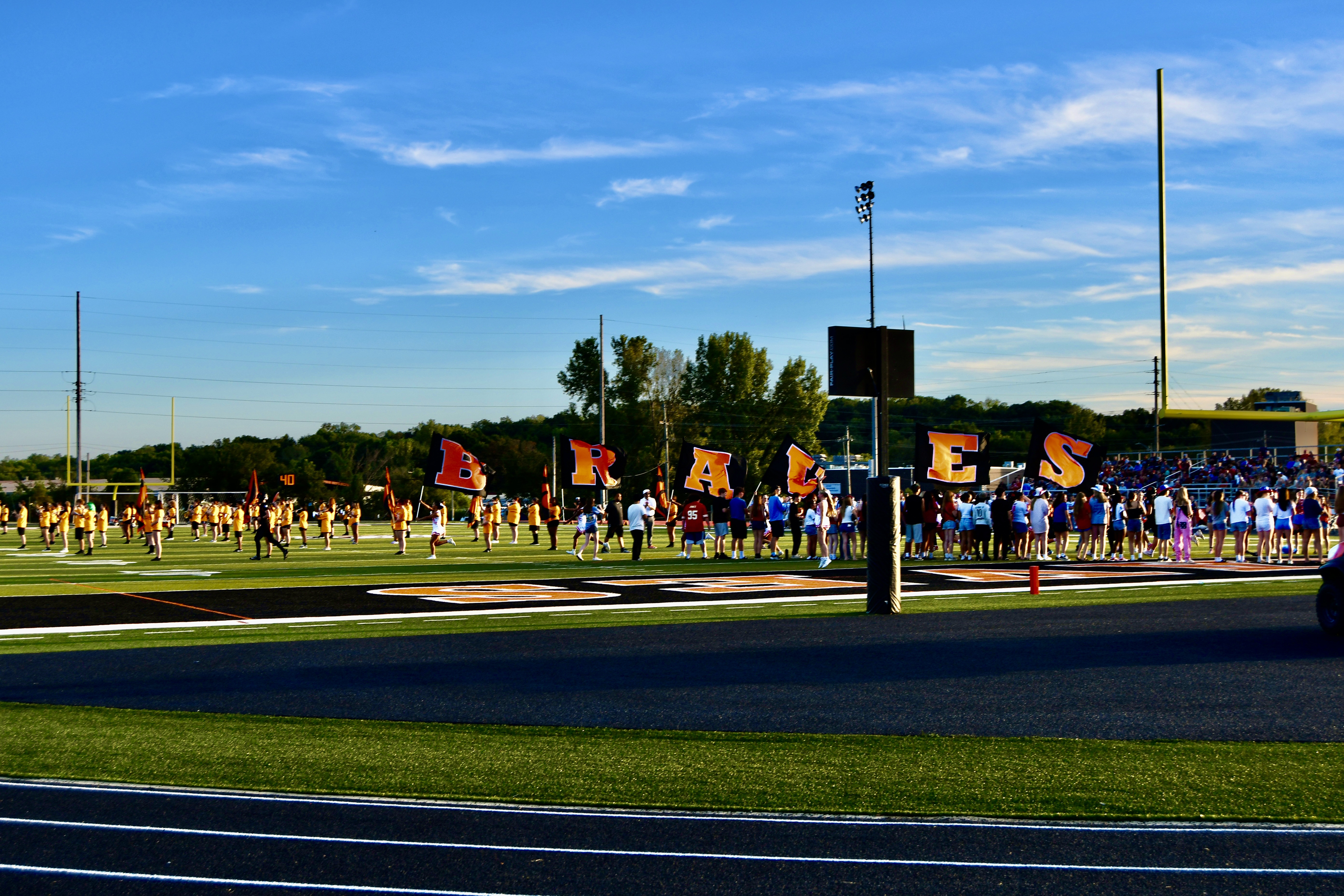 A football field with people gathered, flags, and a sign. Trees and poles in the distance.