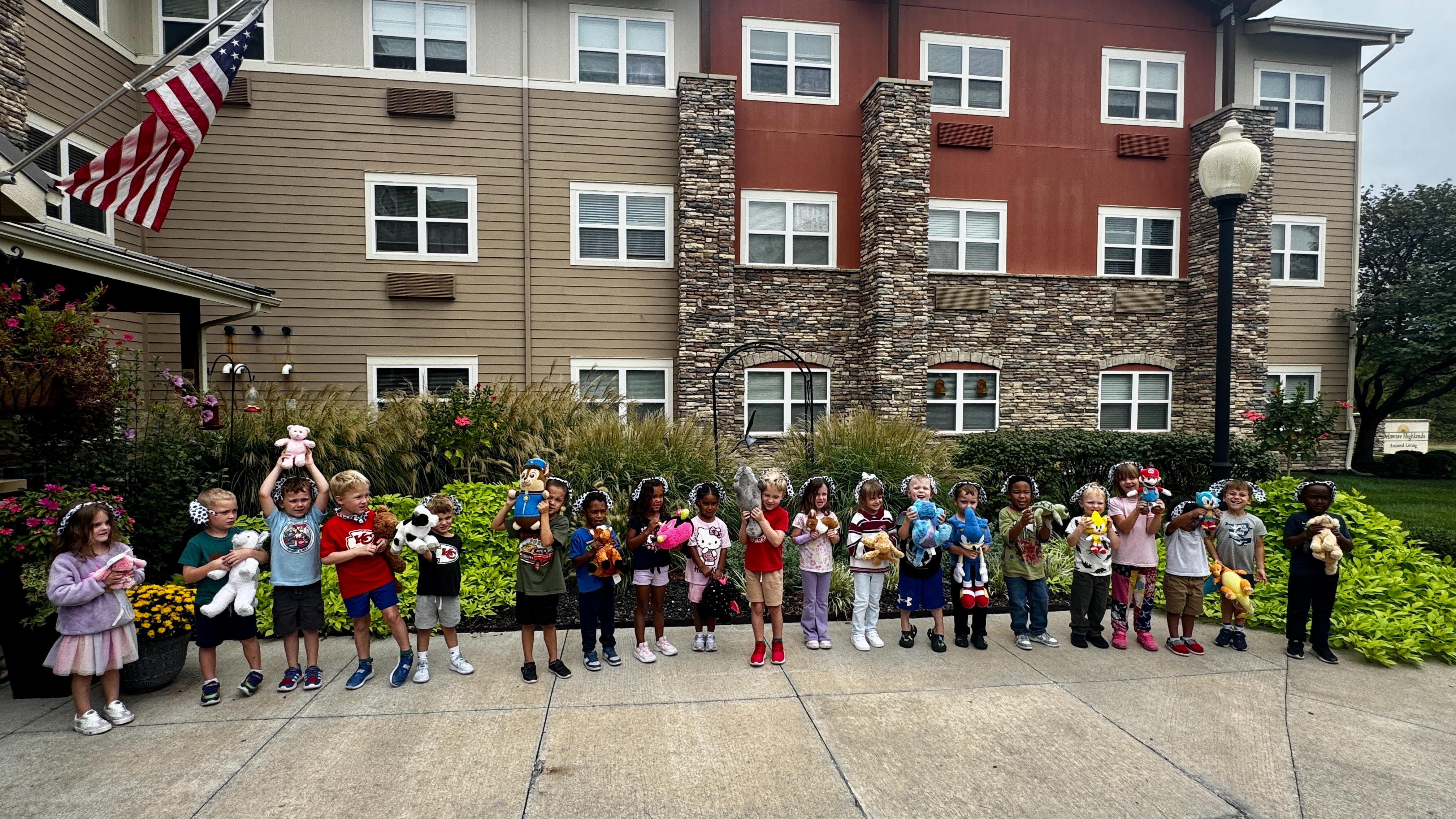 A group of children holding stuffed animals stands outside an apartment complex with a flag.