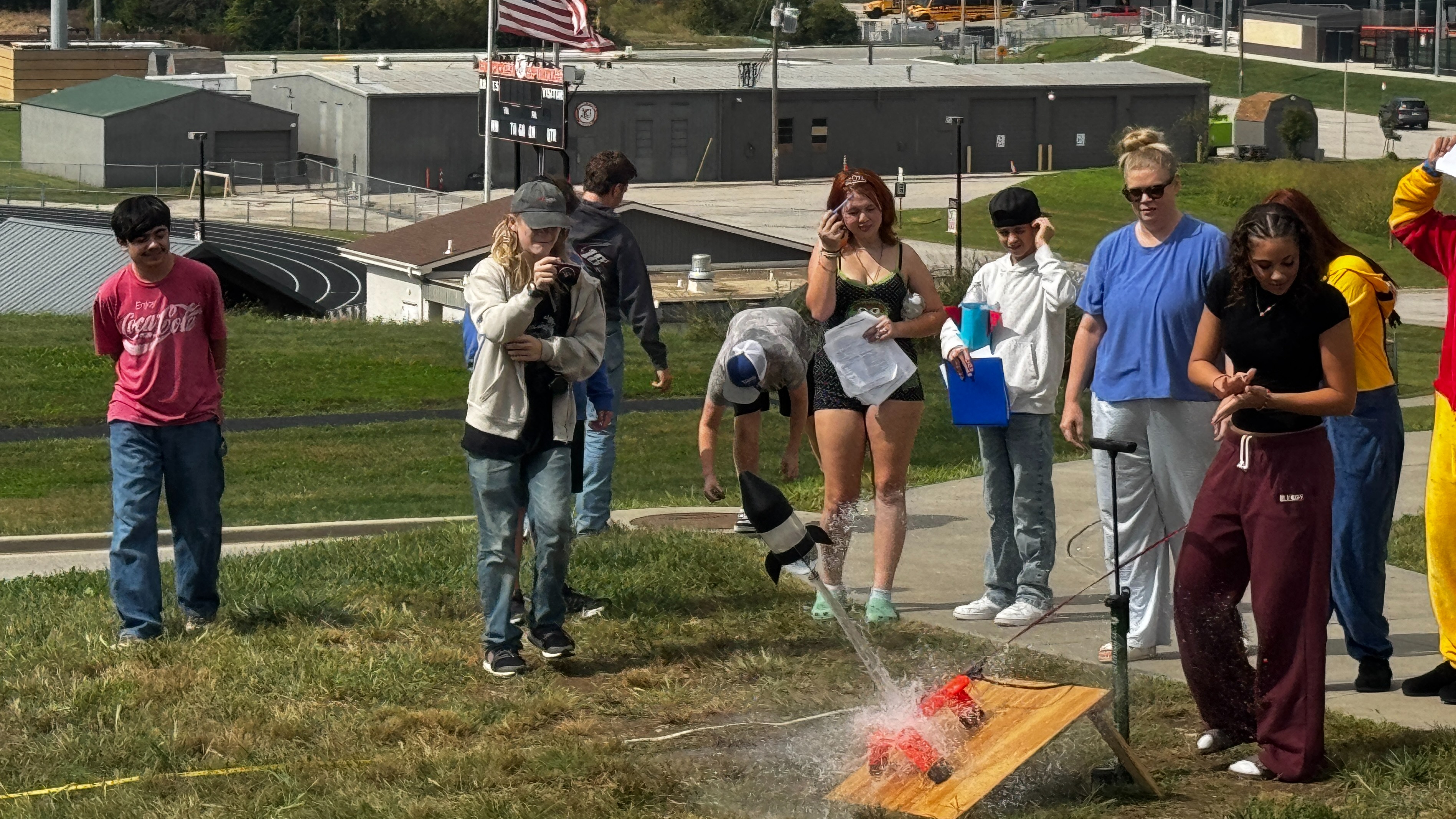 A group of people are gathered in a grassy area with buildings and a flag in the distance.