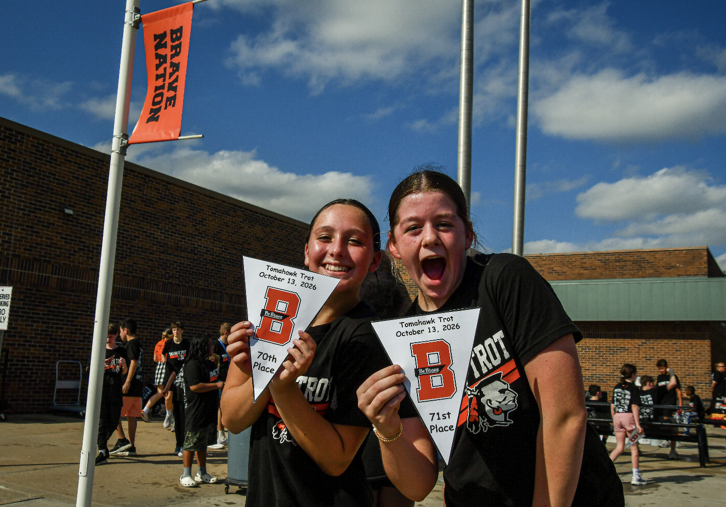 Two students with pennants stand in front of a building with a banner, one holding a pennant with letter B.