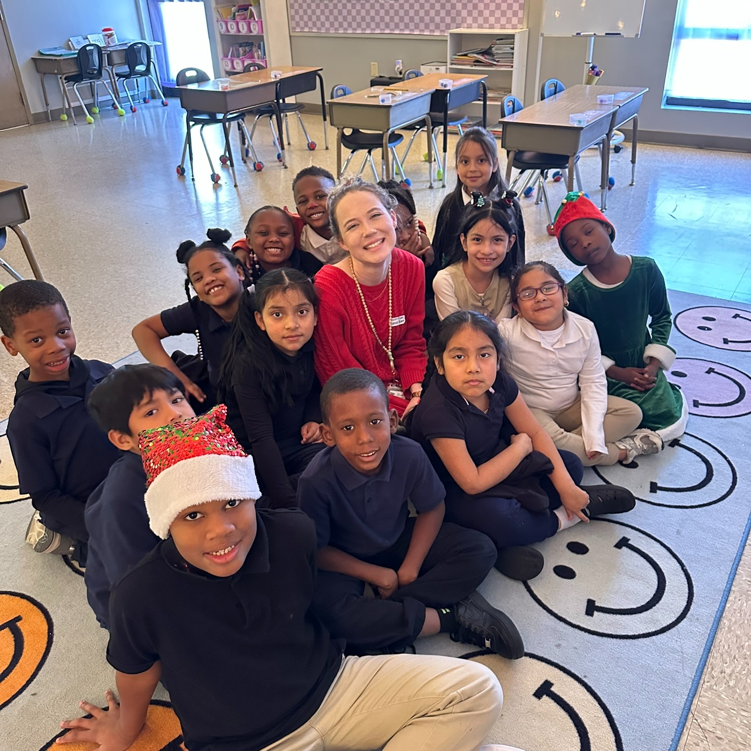 First grade teacher and students smiling while sitting on the floor in the classroom.