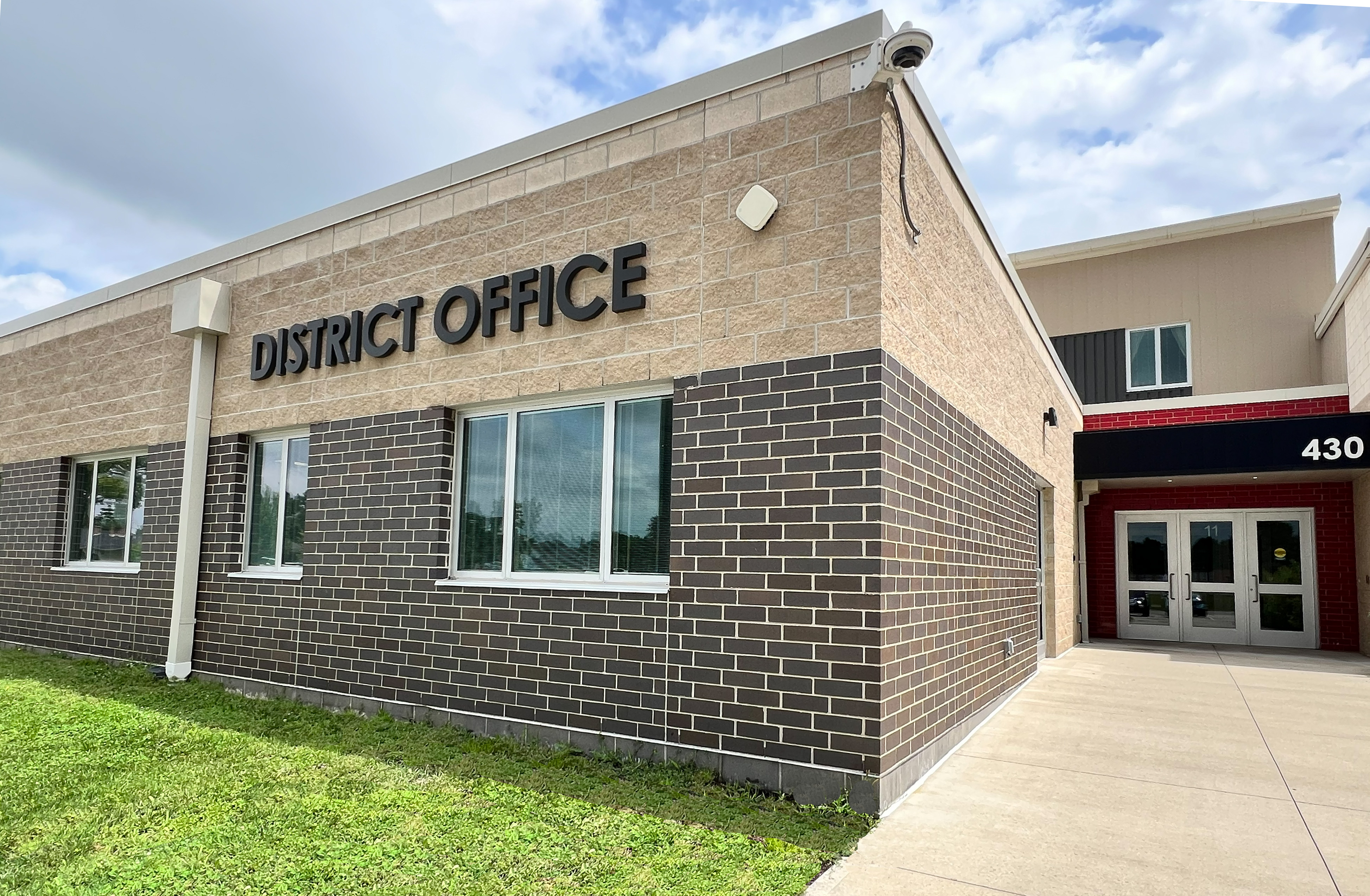 Exterior of a brick building with a sign reading "District Office". Windows, surveillance cameras, and a pathway leading to the entrance.