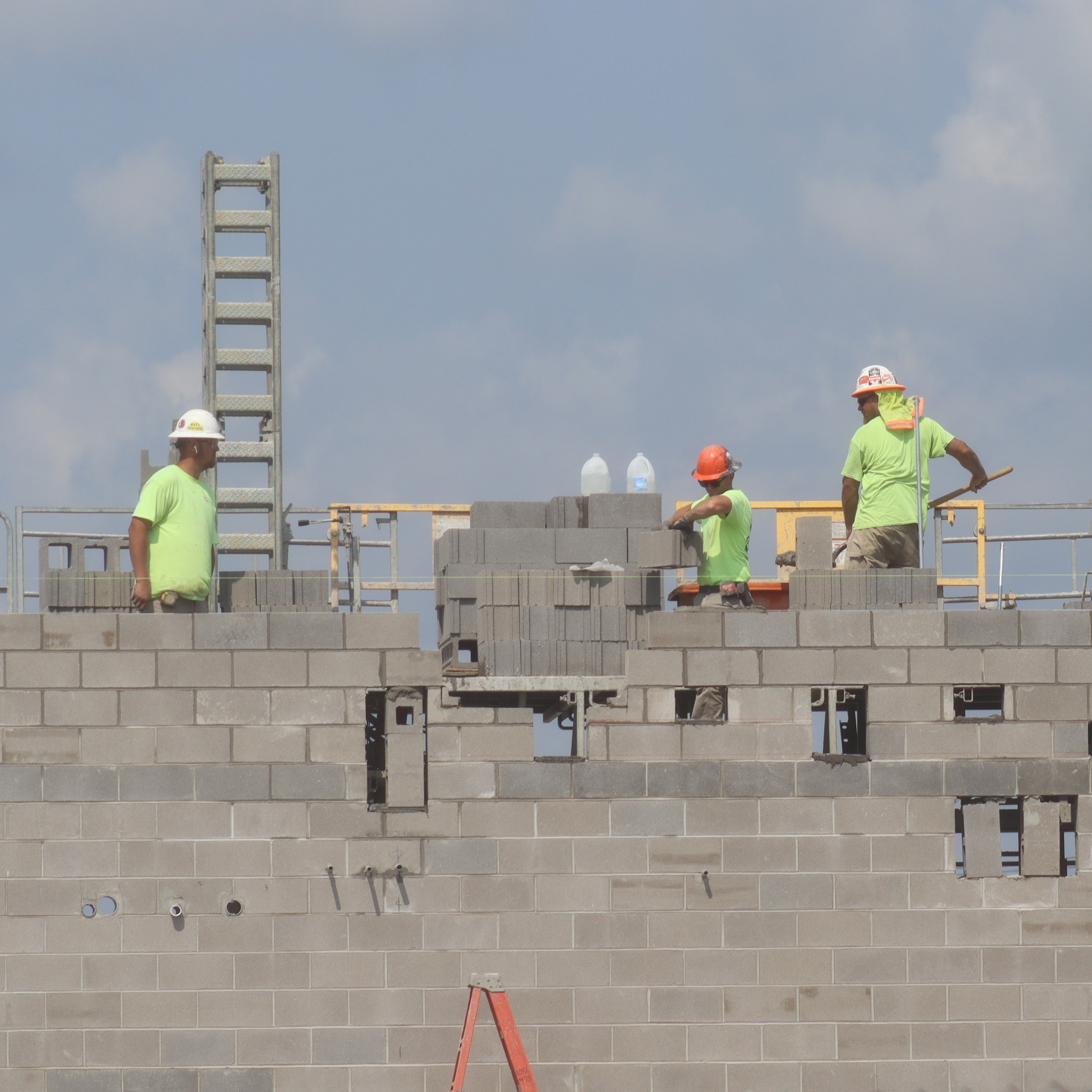 Workers in green shirts and hard hats stand atop a brick structure, with cranes and scaffolding in the background.