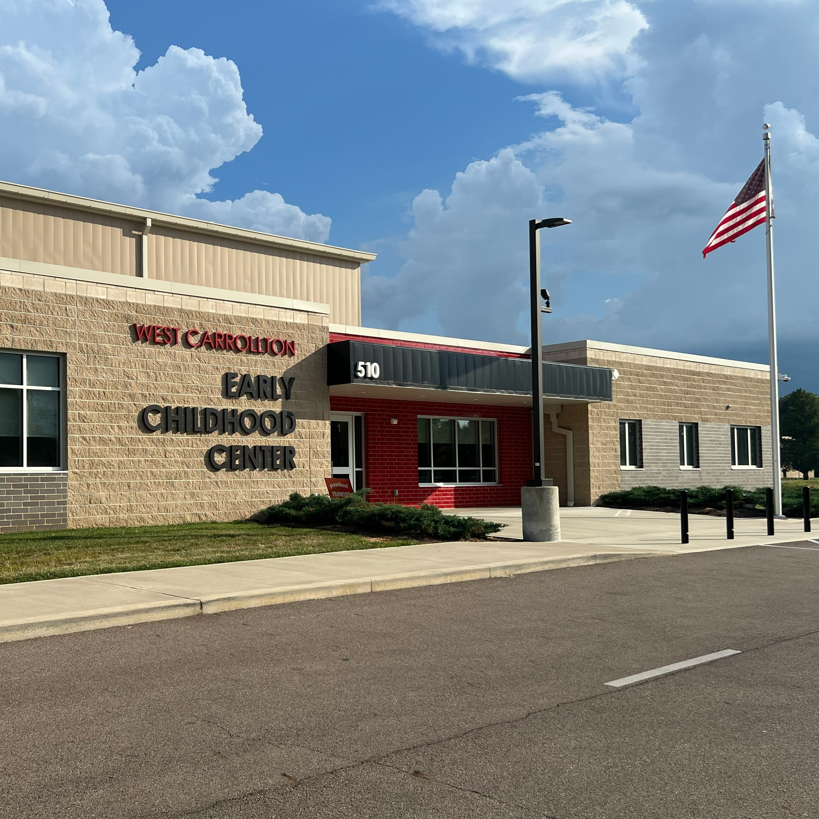 Modern building with large windows and red door. Text reads "Early Childhood Center". American flag on a pole. Empty road in front.