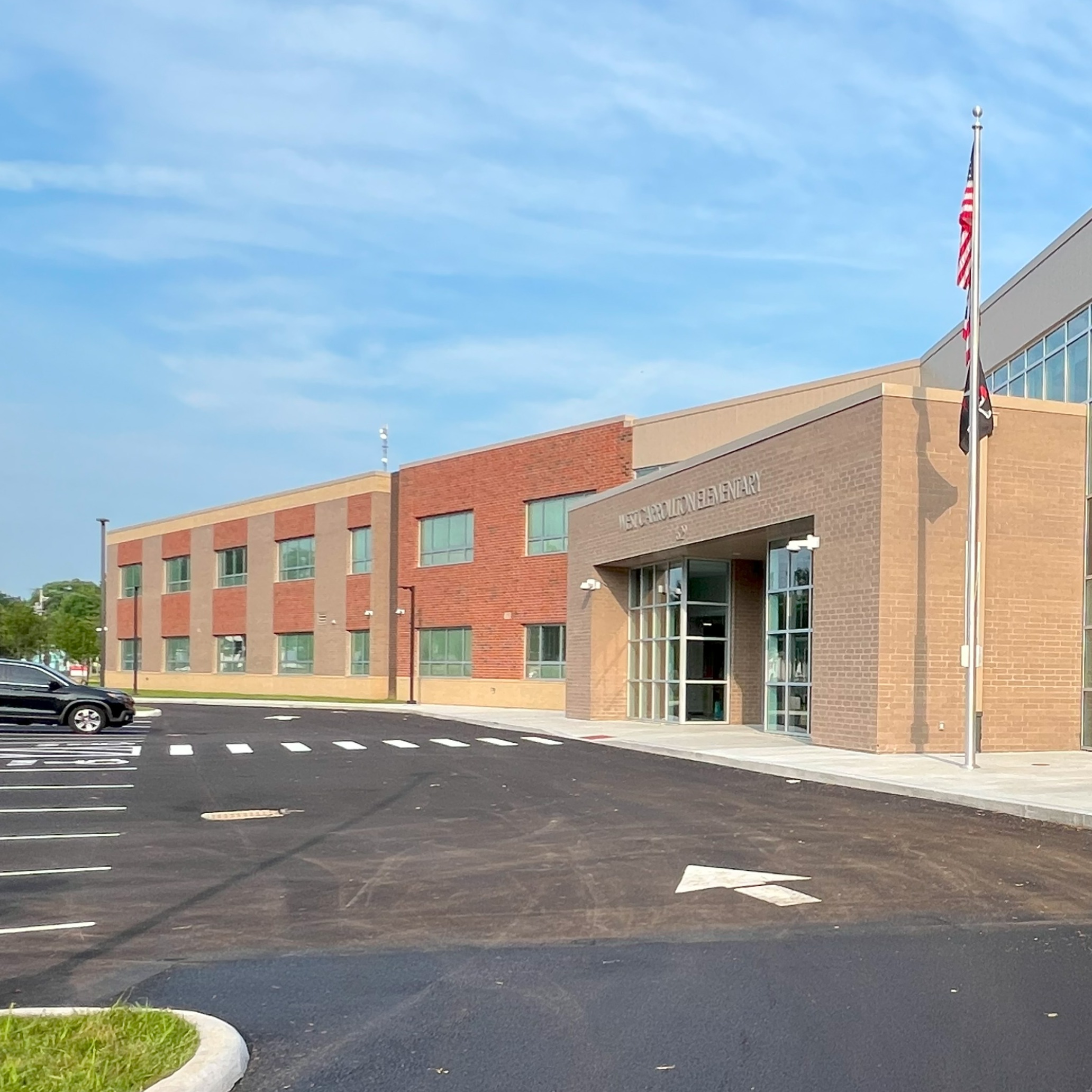 Exterior of a two-story brick building with glass windows. A flag is visible on the building's left. An empty road leads to the front entrance.