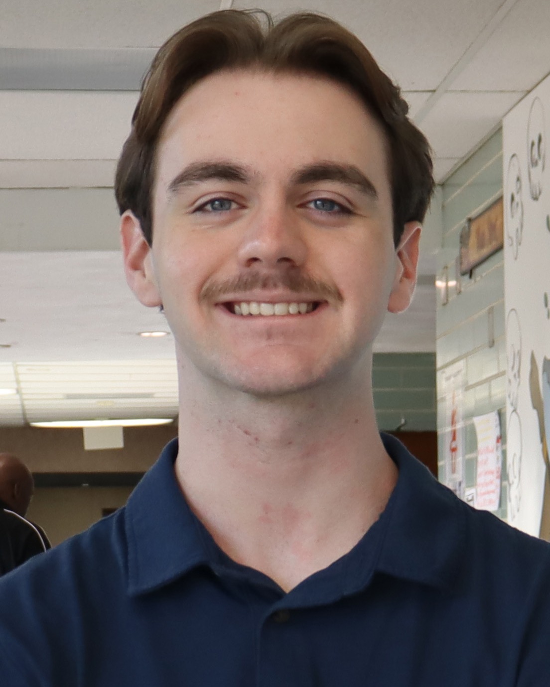 A man with short brown hair and a mustache smiles at the camera. He wears a dark blue collared shirt.