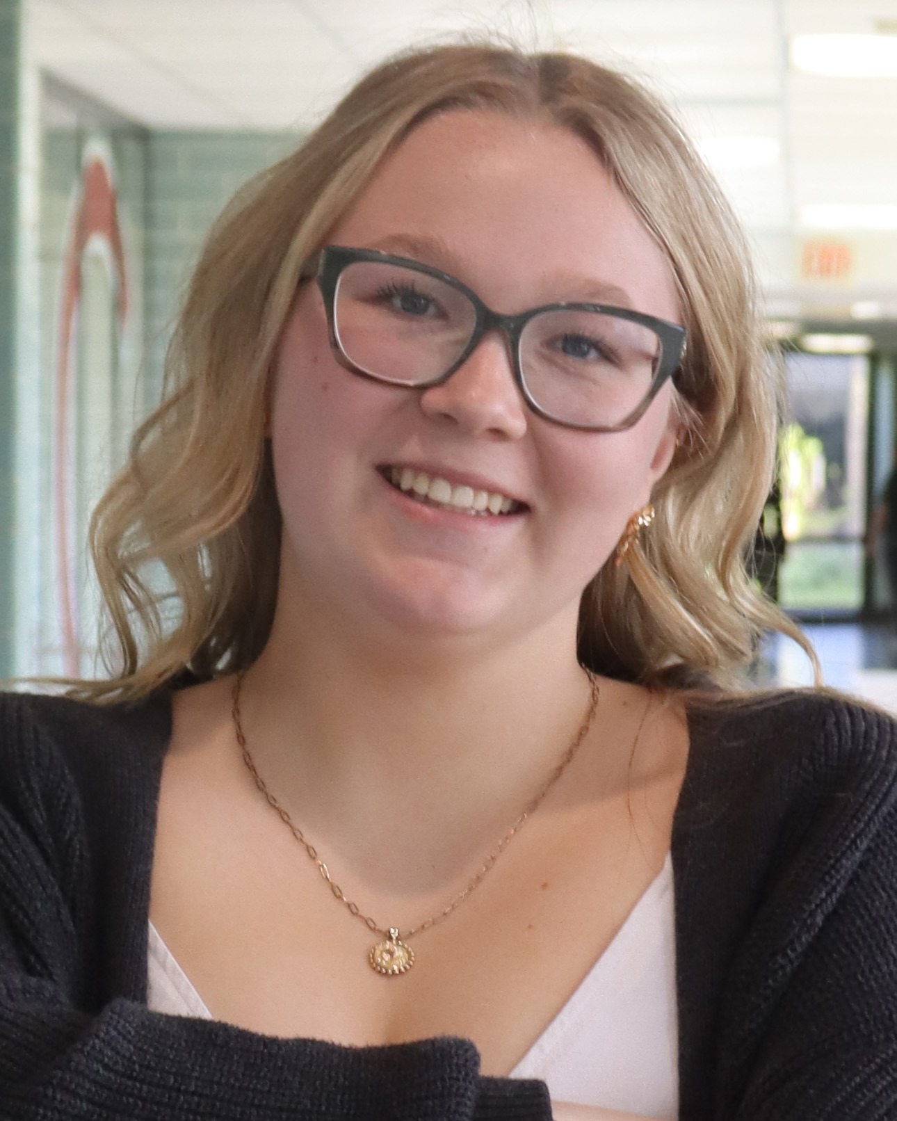 Blonde woman with glasses smiling. She wears a black cardigan over a white top and a gold necklace with a circular pendant.