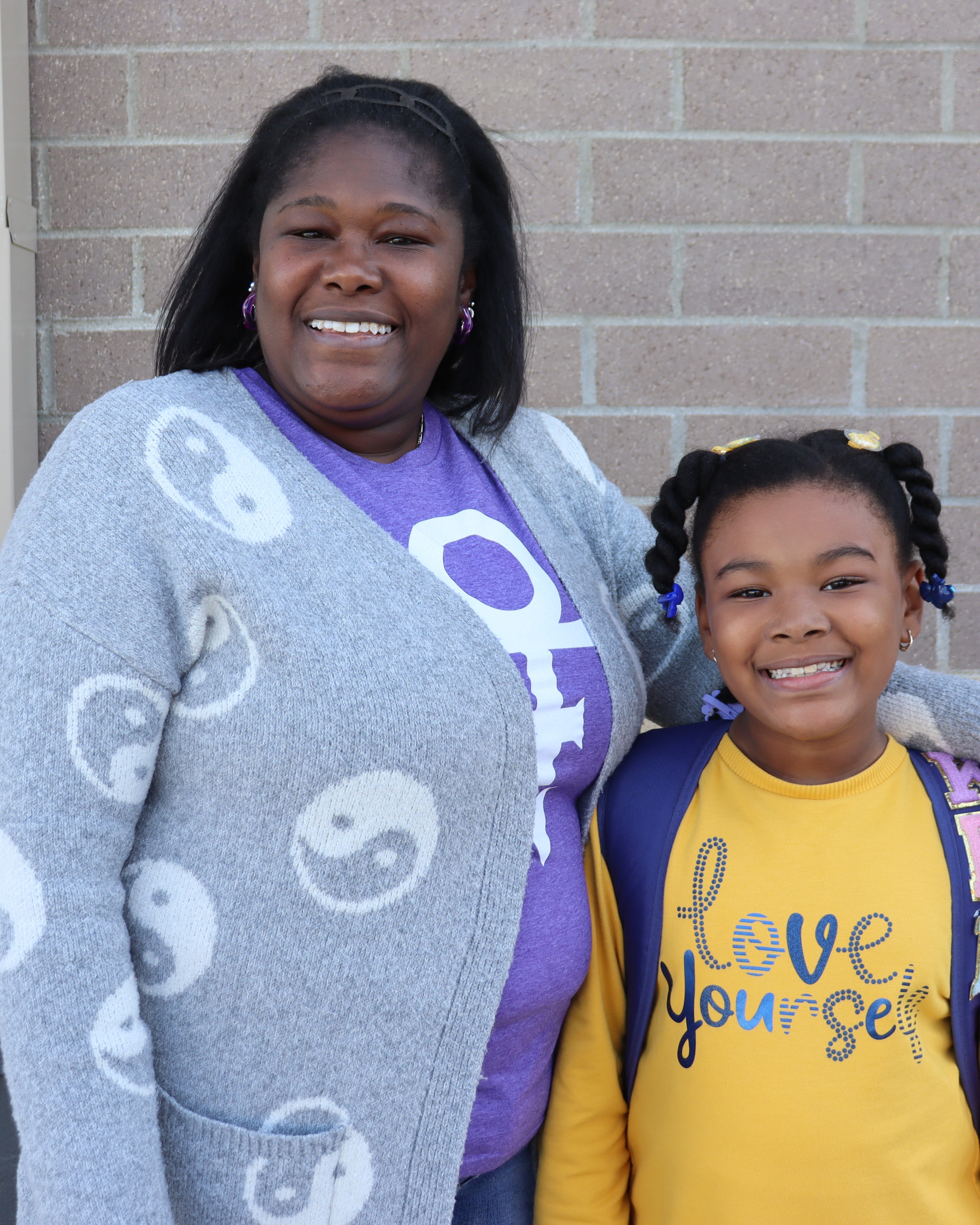 A woman with a gray cardigan and a young girl in a yellow shirt smile outside a brick building.
