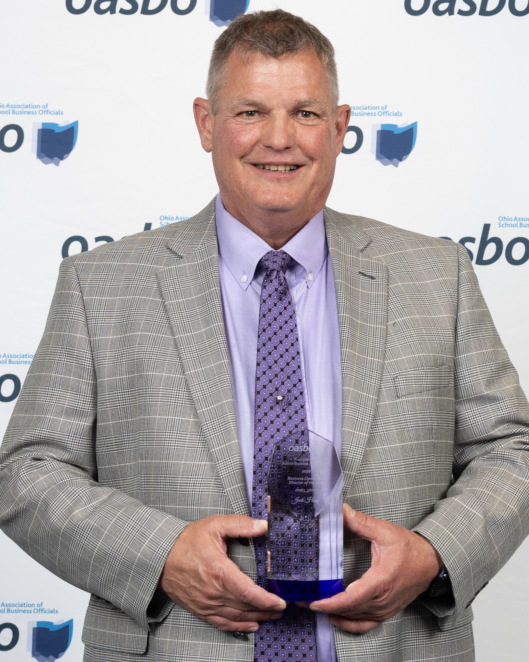 A man in a gray suit and purple tie stands in front of a wall with logos, holding an award.