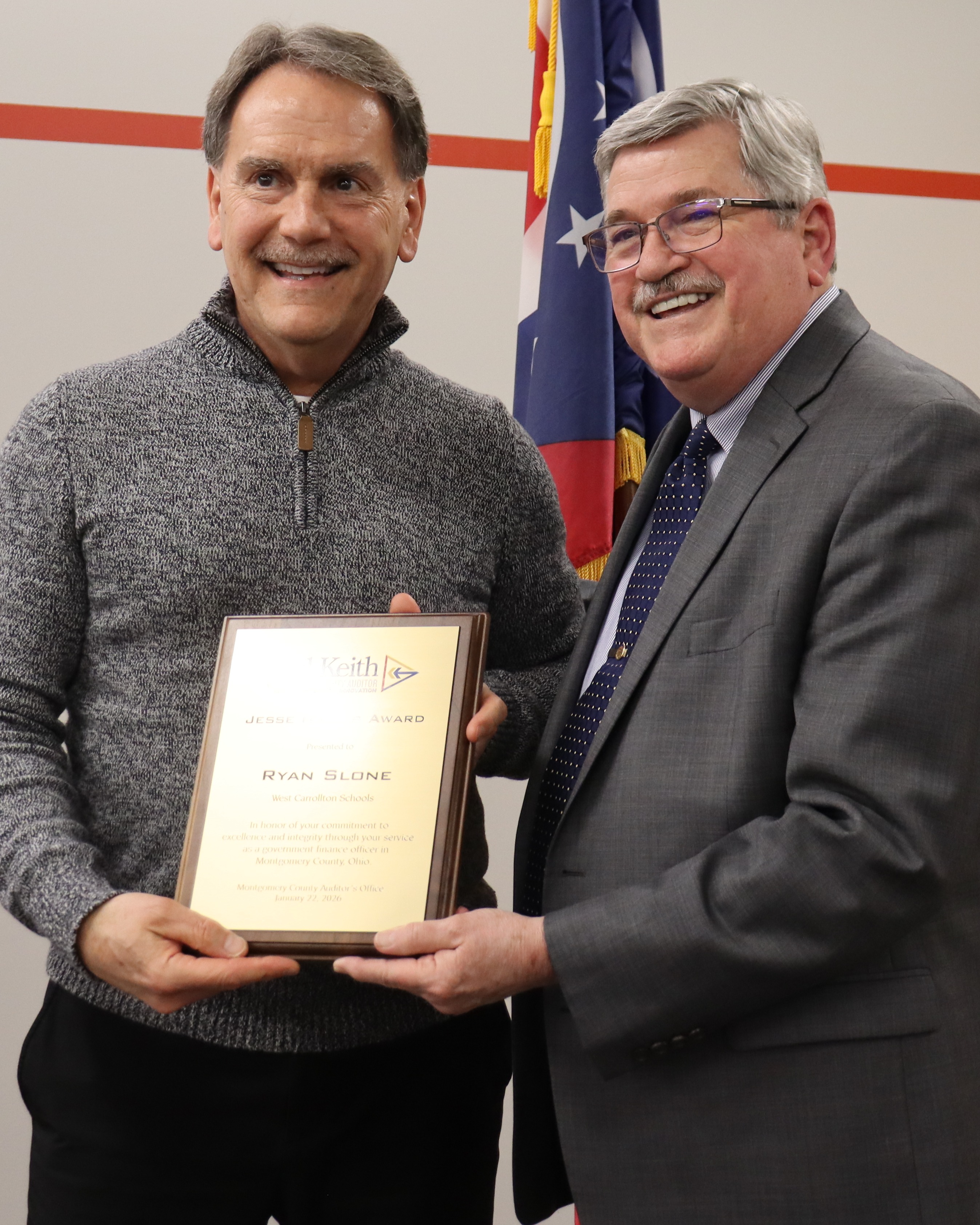 Two men standing in a room, one holding a framed certificate. One wears a gray sweater, the other a gray suit. Behind them is a flag and a wall.