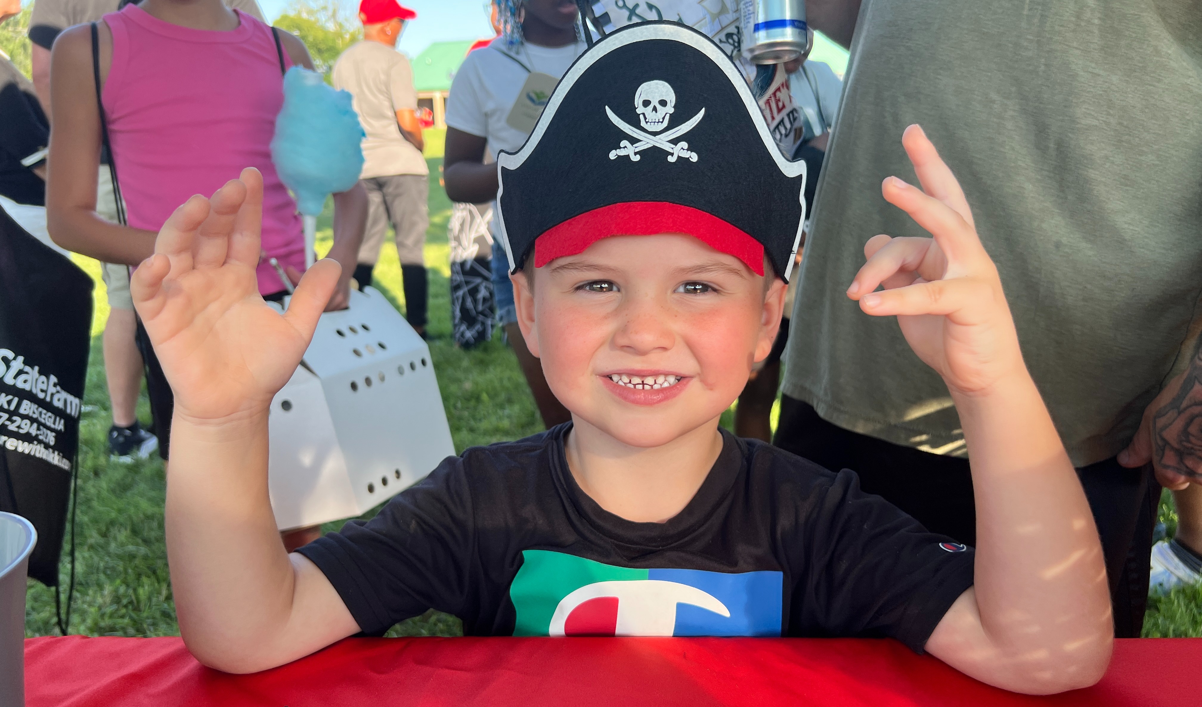 A young boy in a pirate hat sits at a table, smiling and making a peace sign.