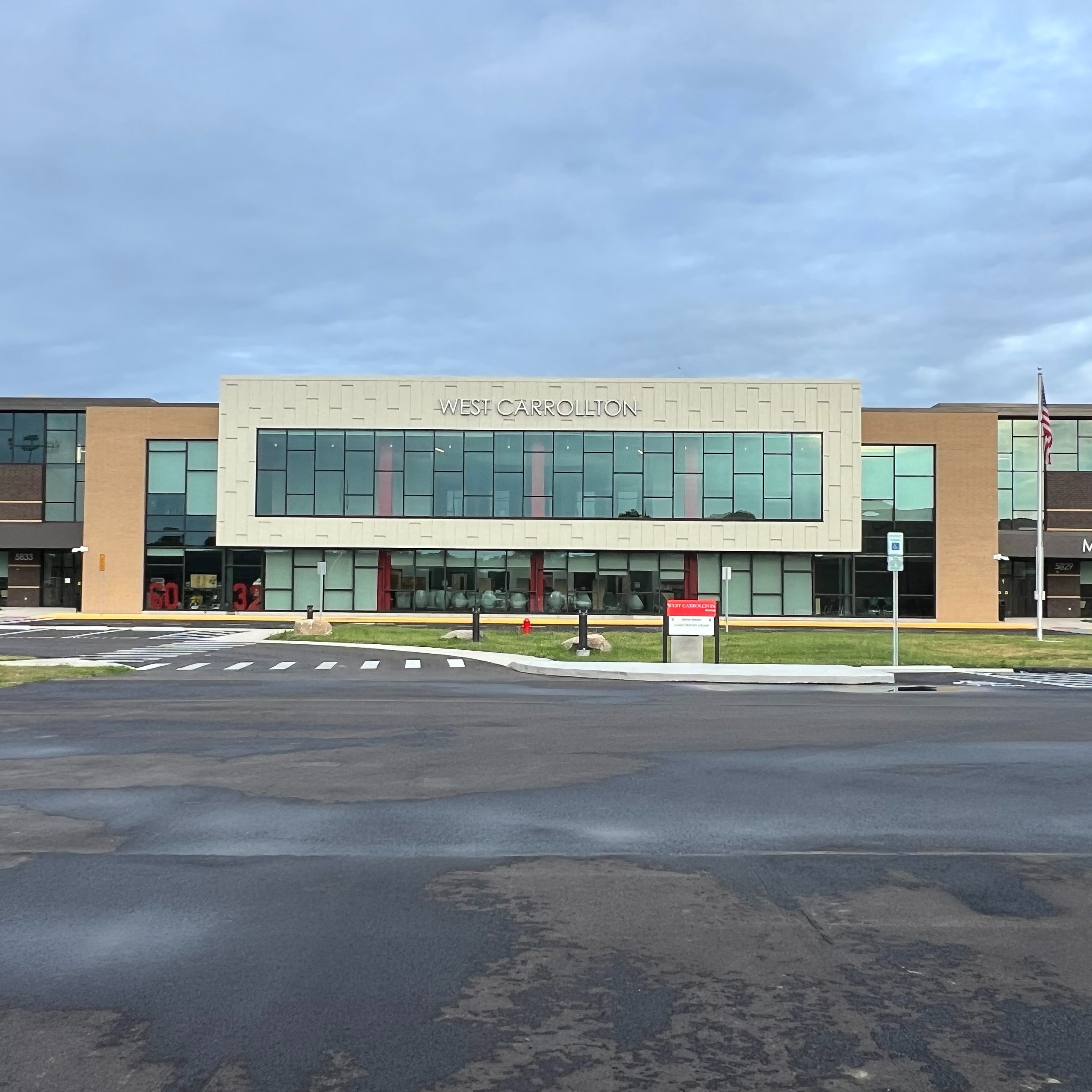 An empty school parking lot in front of a large building with glass windows. A flag pole stands on the right.
