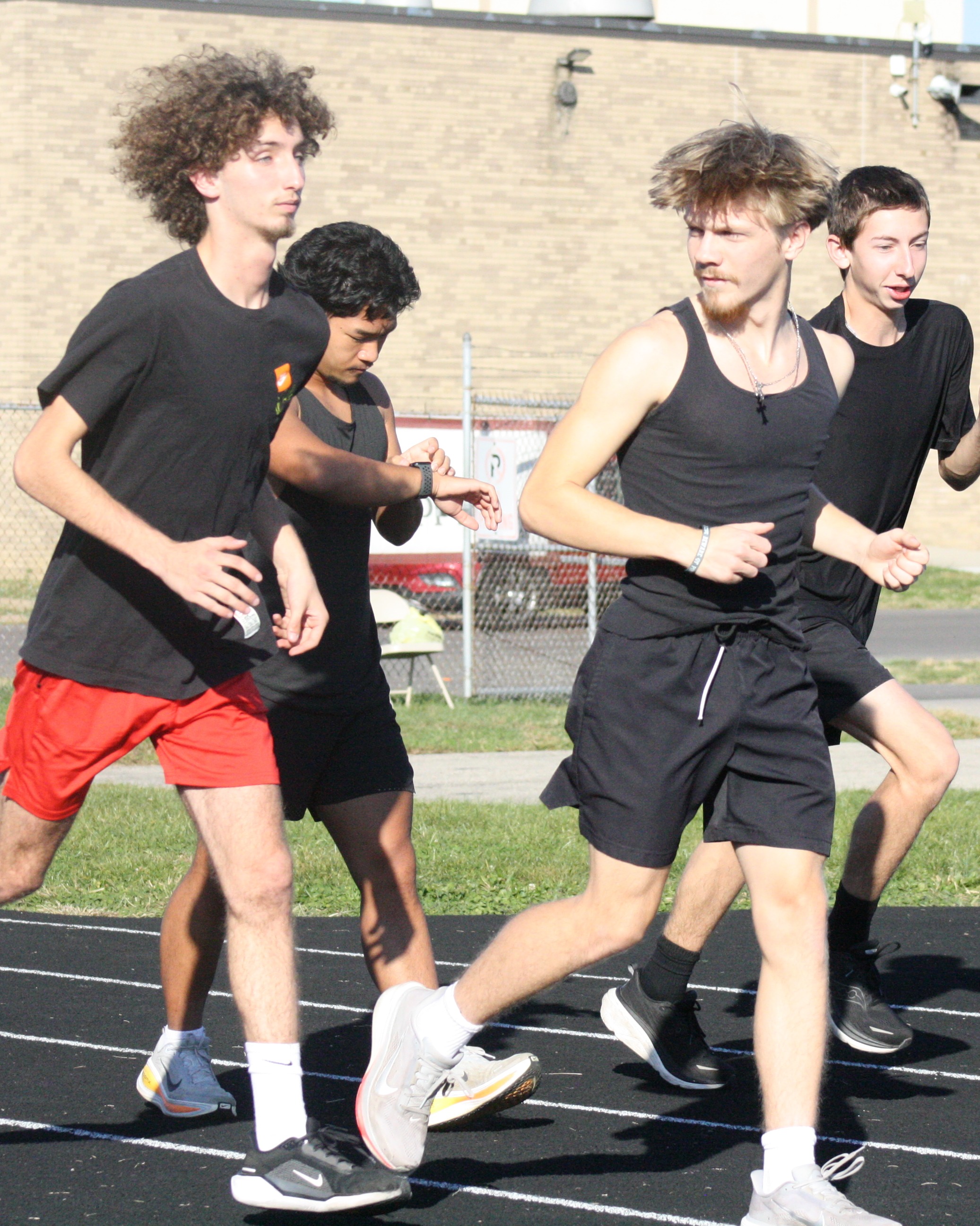 Four boys in athletic wear run on a track, their shoes making imprints in the dirt.