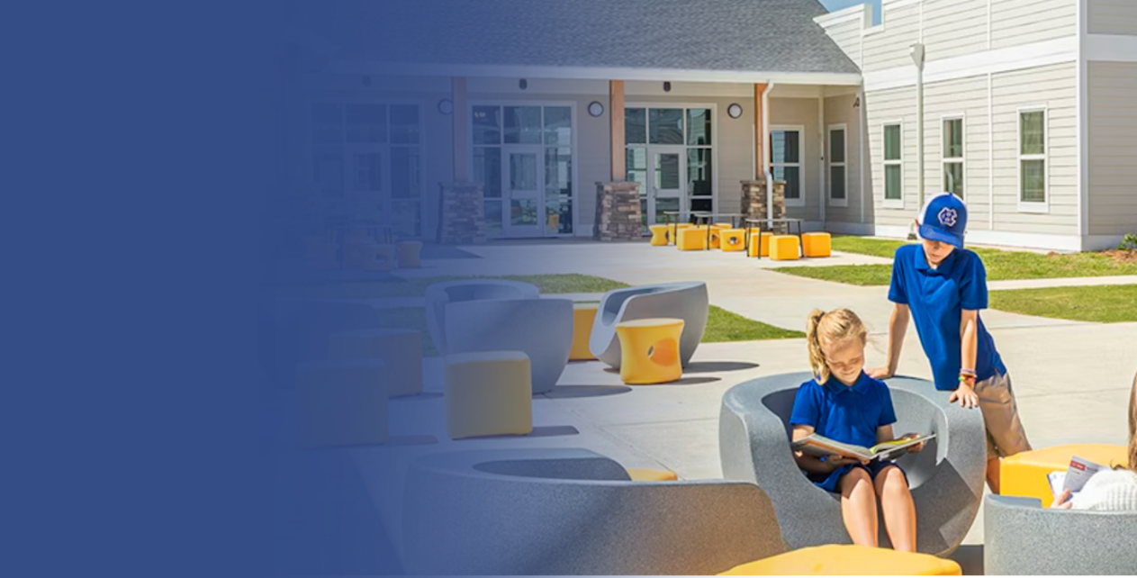 Children seated in outdoor furniture in front of a building. One reads a book. Sunny, outdoor scene.