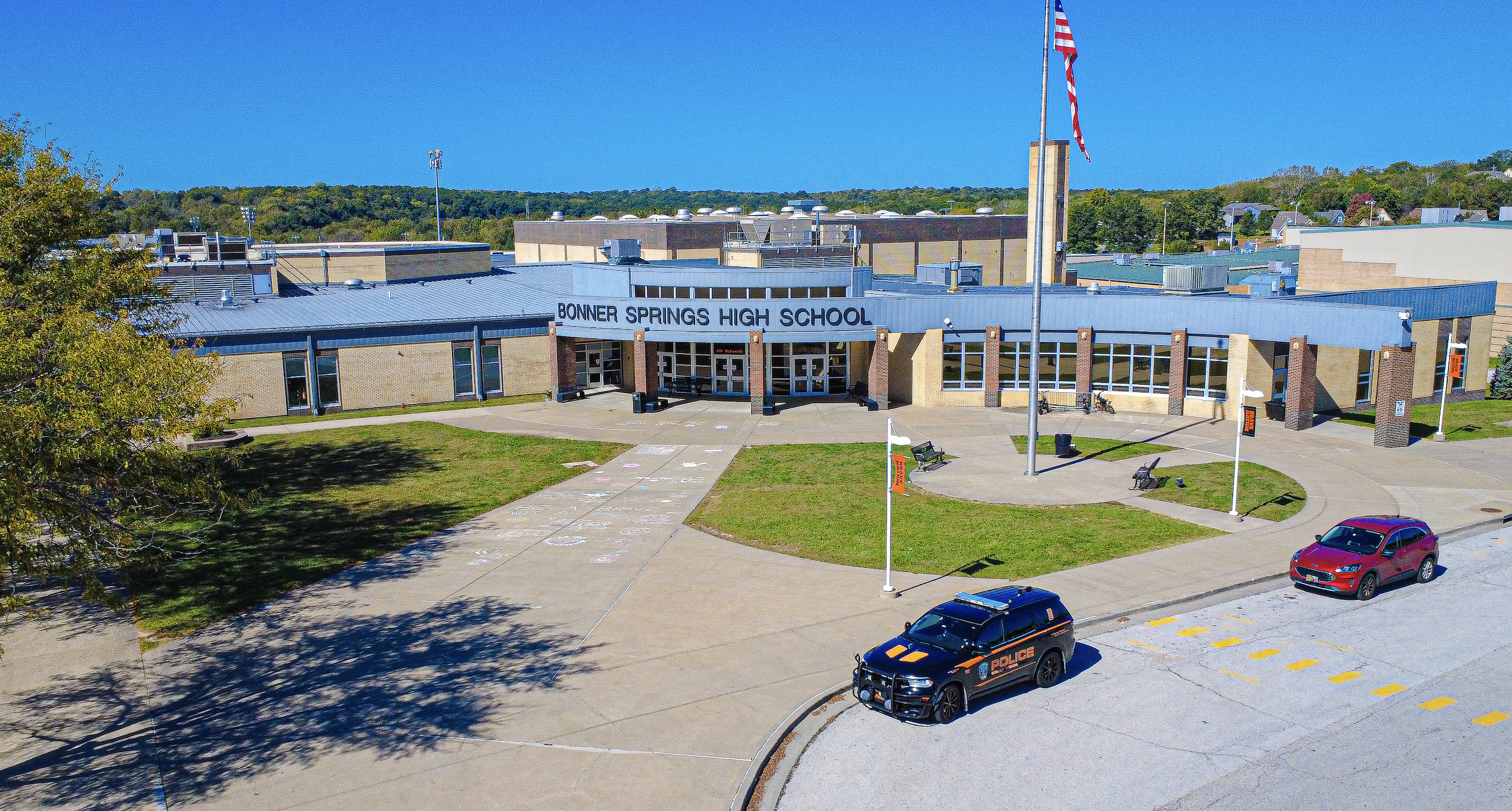 A school building with a flag on top has cars parked in front, one black, one red.