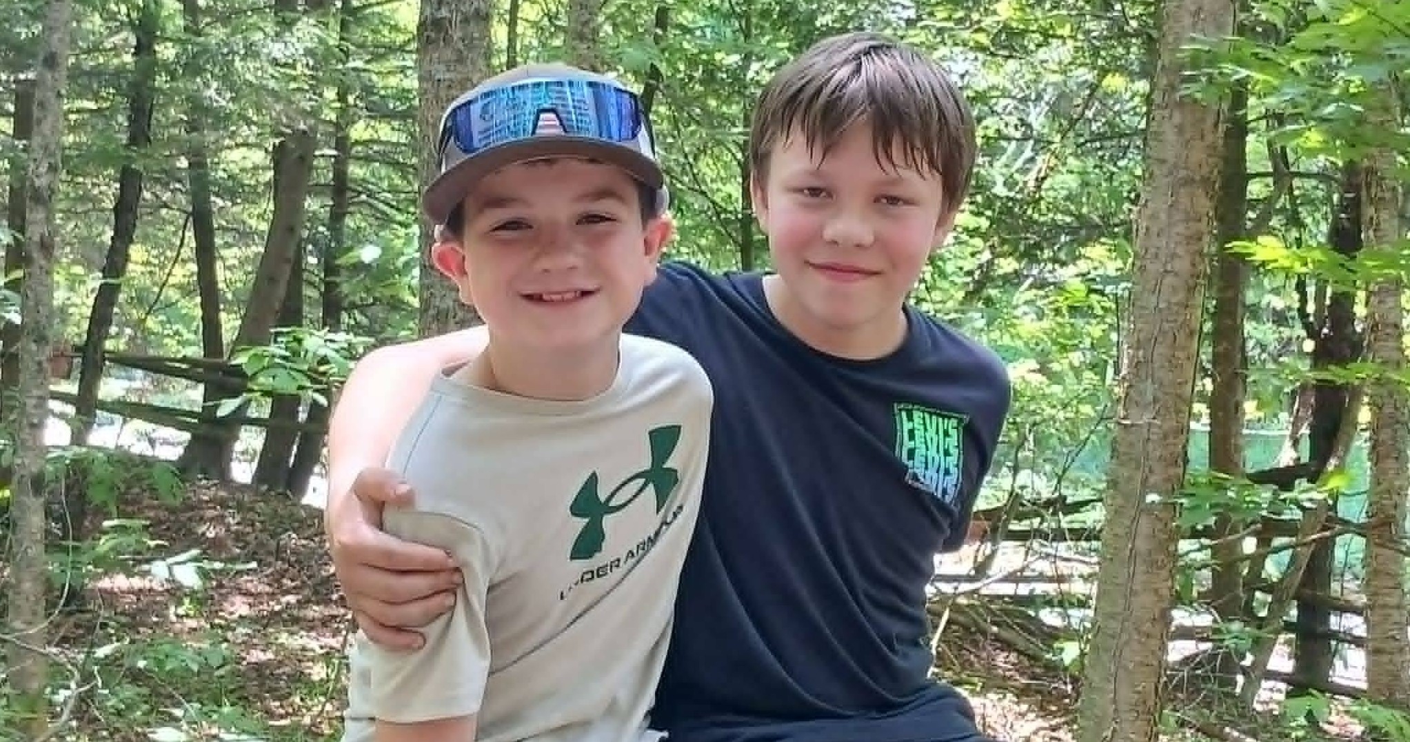 Two students smile for photo in wooded area, one has an arm around the other.