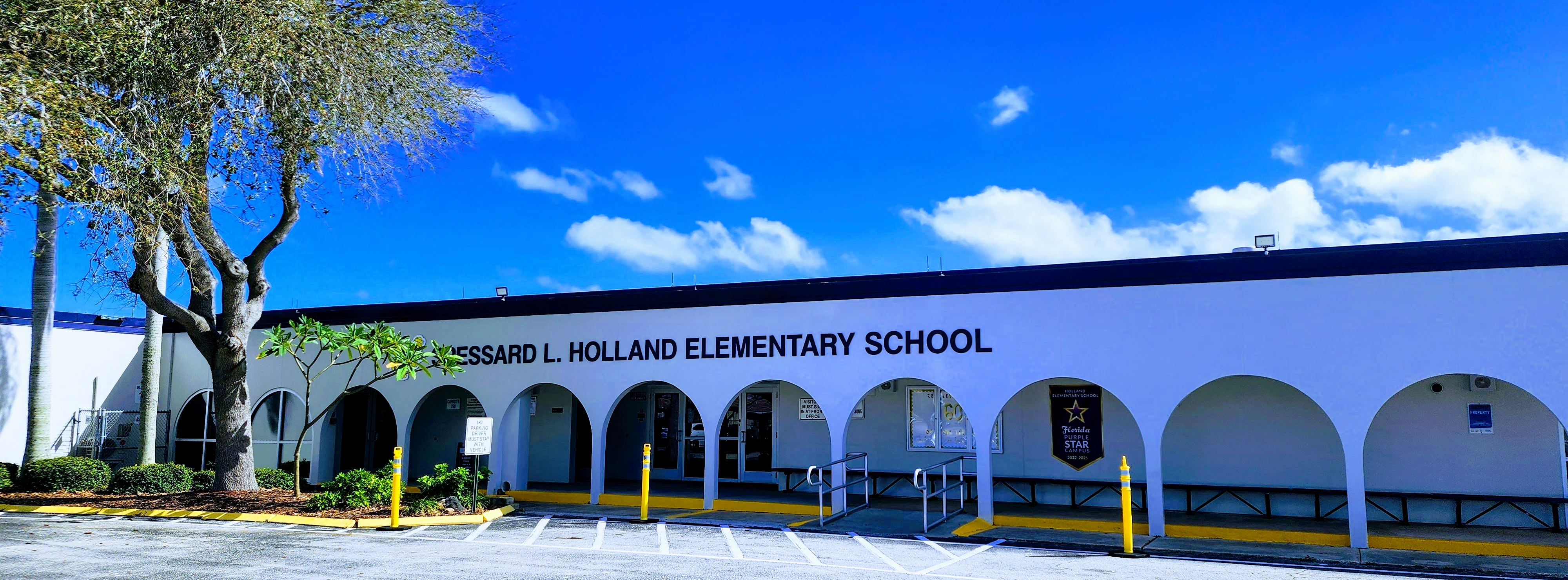 Holland Elementary front of building , white buiilding with blue trim, Spessard L. Holland Elementary School sign. Large tree to the left of the building.