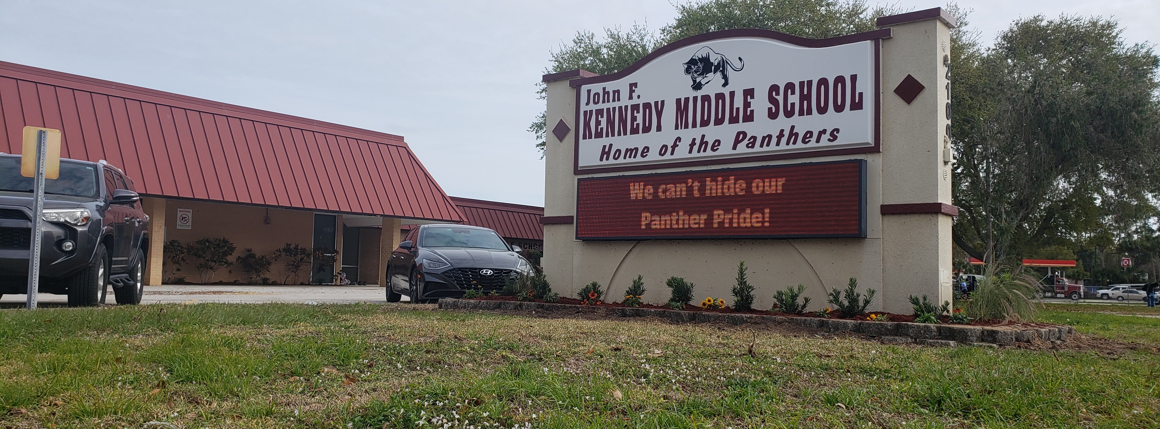 Picture of the front of Kennedy Middle school with a sign that says"John F. Kennedy Middle School Home of the Panthers. Below that  there is an electoric sign that says "We cant hide our panther pride!" 