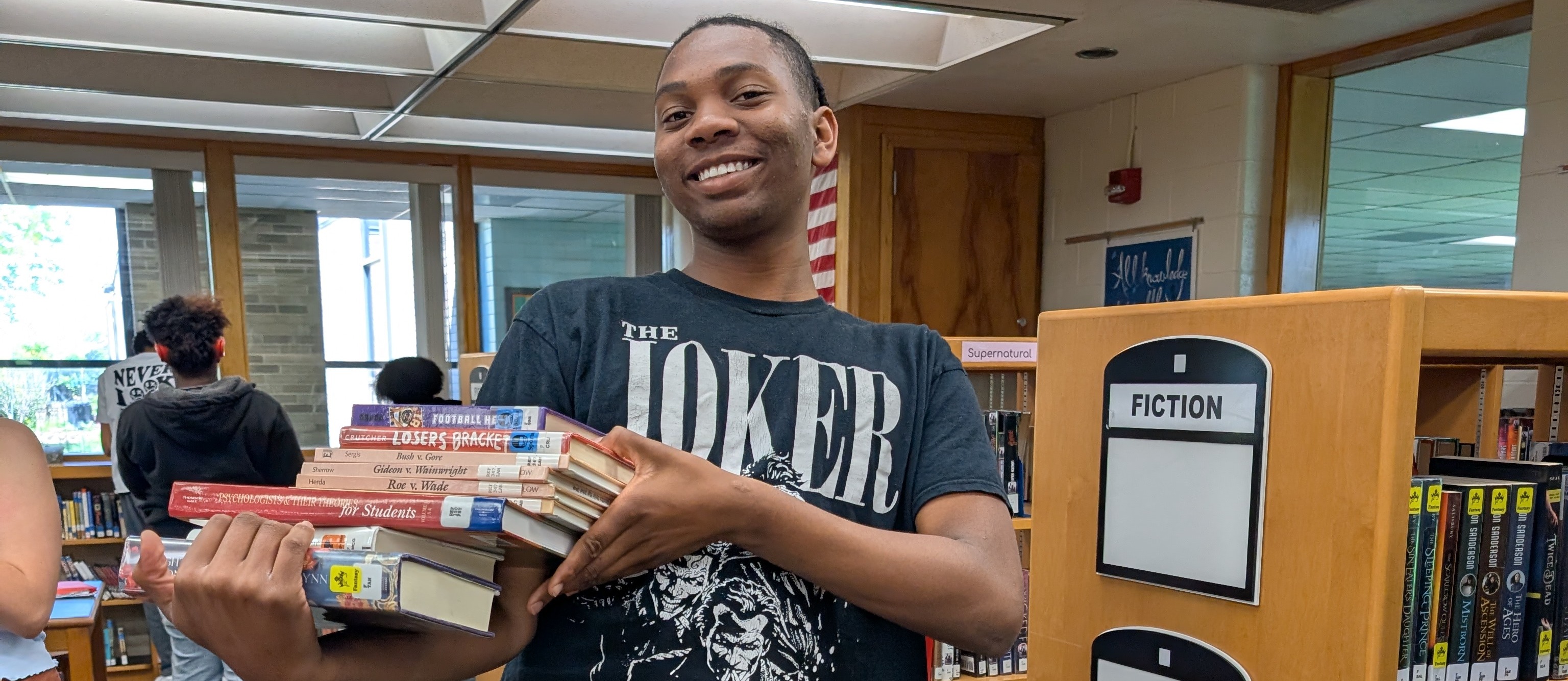 A person holds a stack of books in a library with shelves and other people in the background.