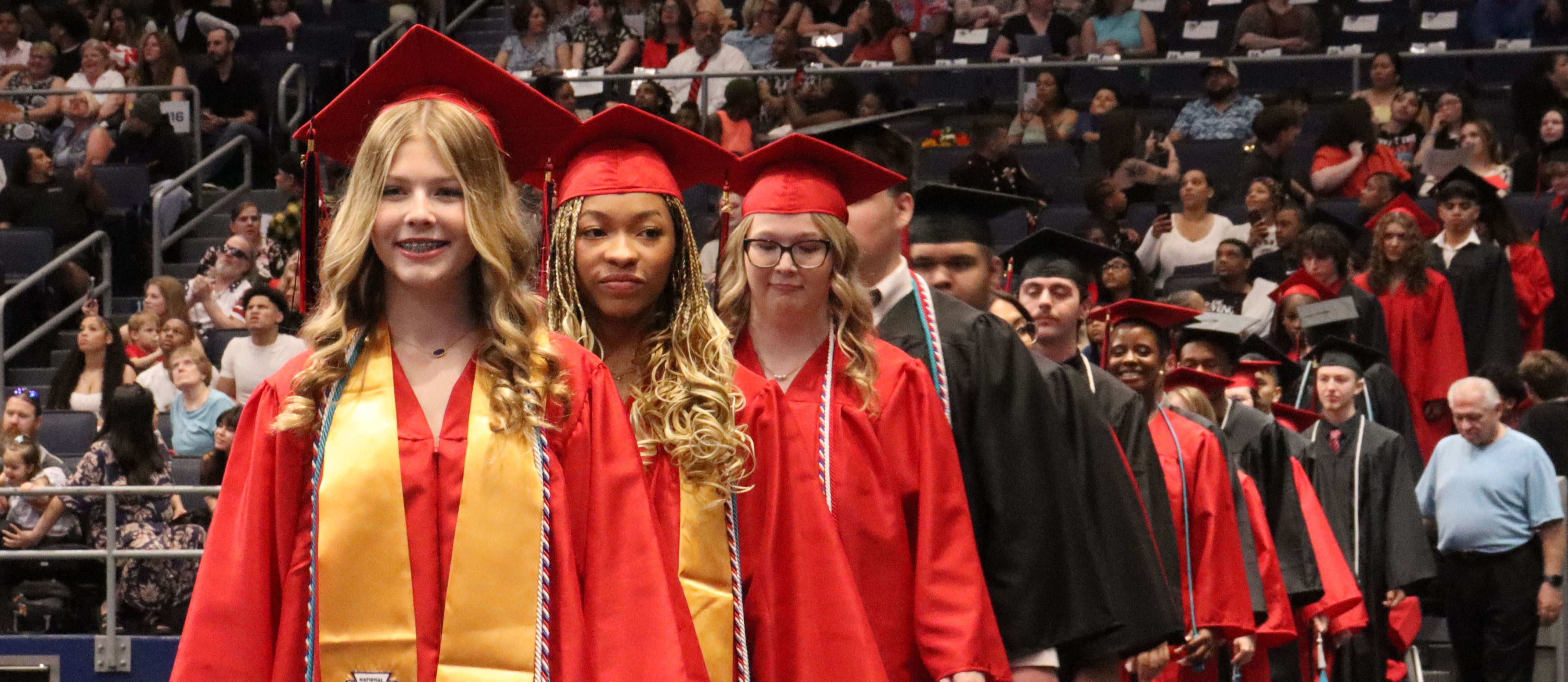 Graduates in red gowns and caps walk on a stage; a crowd watches from the stands.