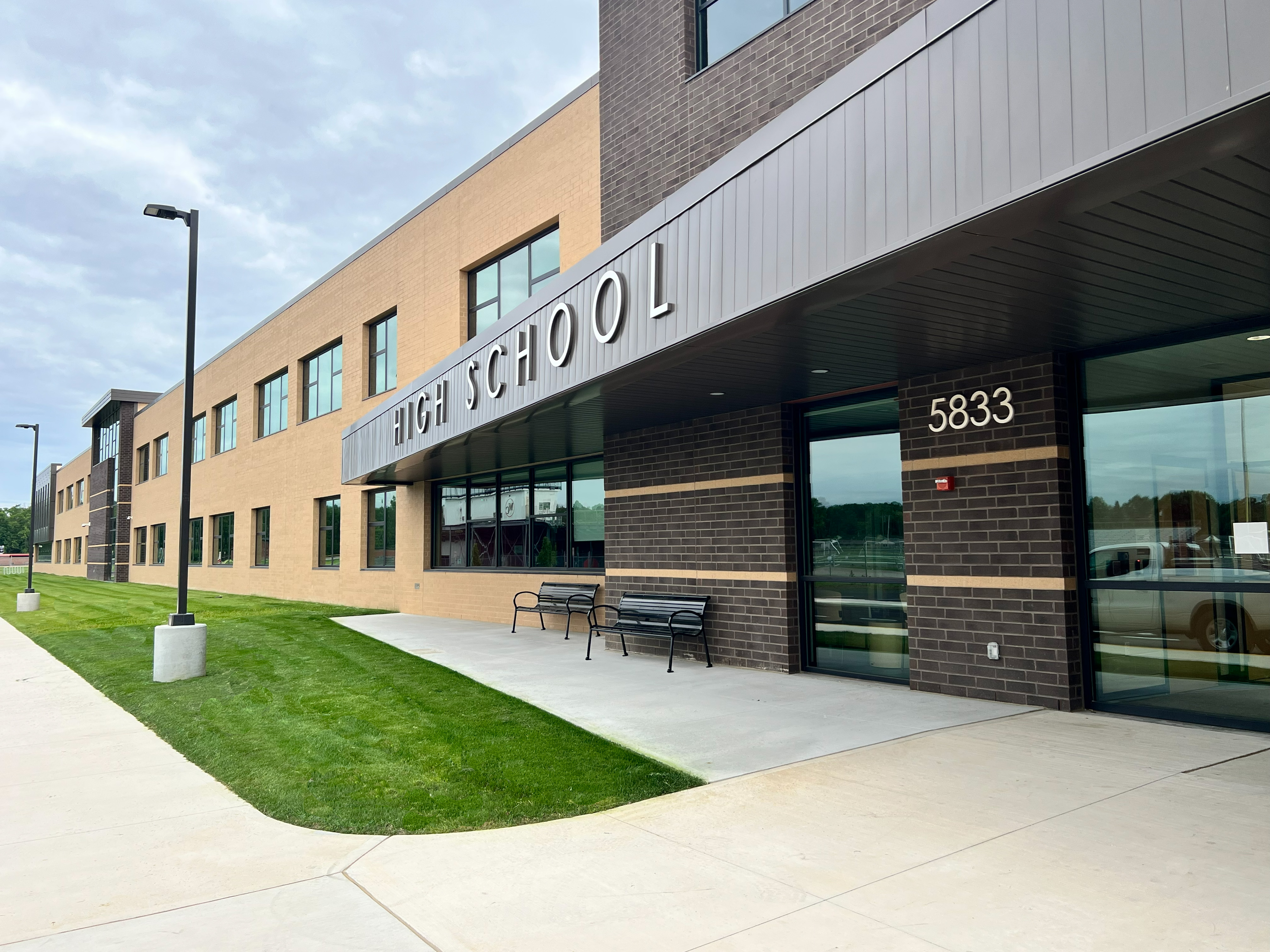 Modern school building entrance with a paved path, green grass, benches, a streetlight, and a sign reading "High School 5833."