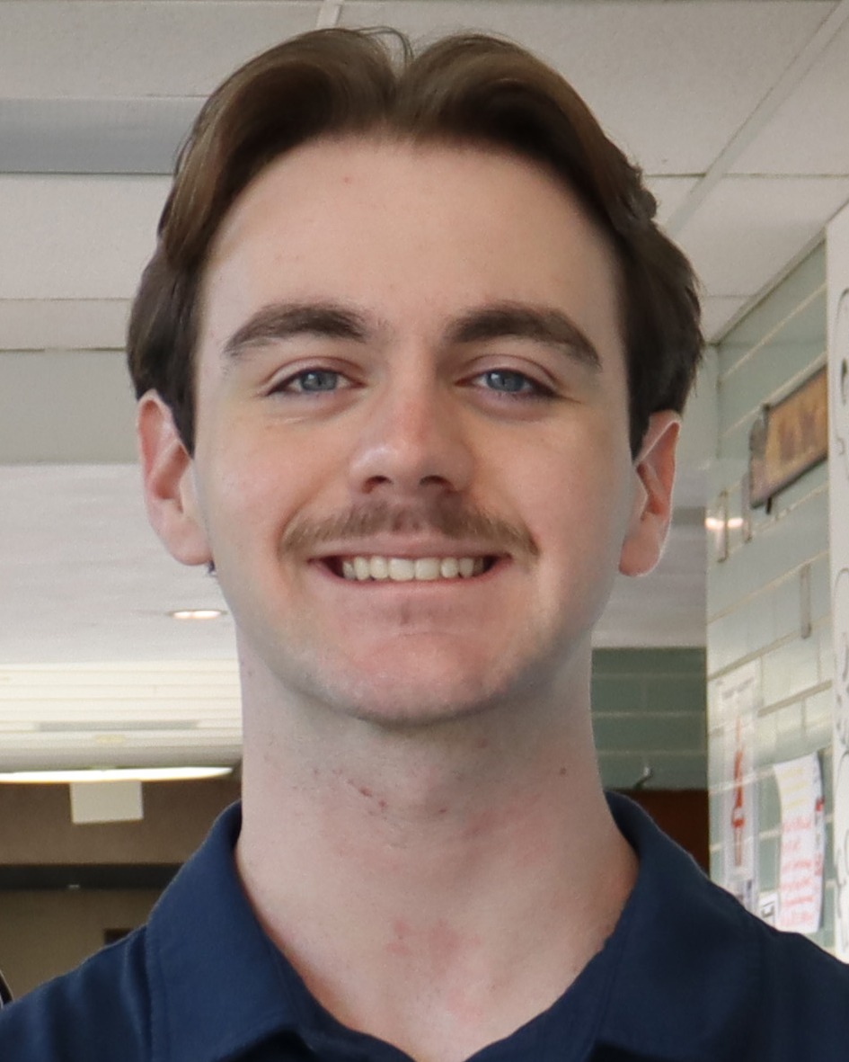 A man with brown hair and a mustache smiles in a corridor. He wears a navy blue polo shirt.