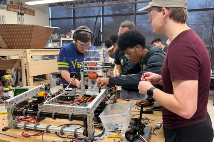 Students in a workshop work on a metal structure with wires and tools on a workbench.
