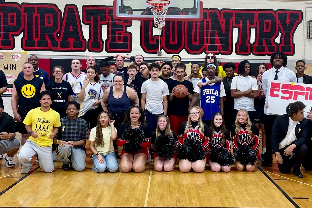 A group of people in a gym posing for a photo, with a basketball hoop, pirate country sign, and wooden floor.