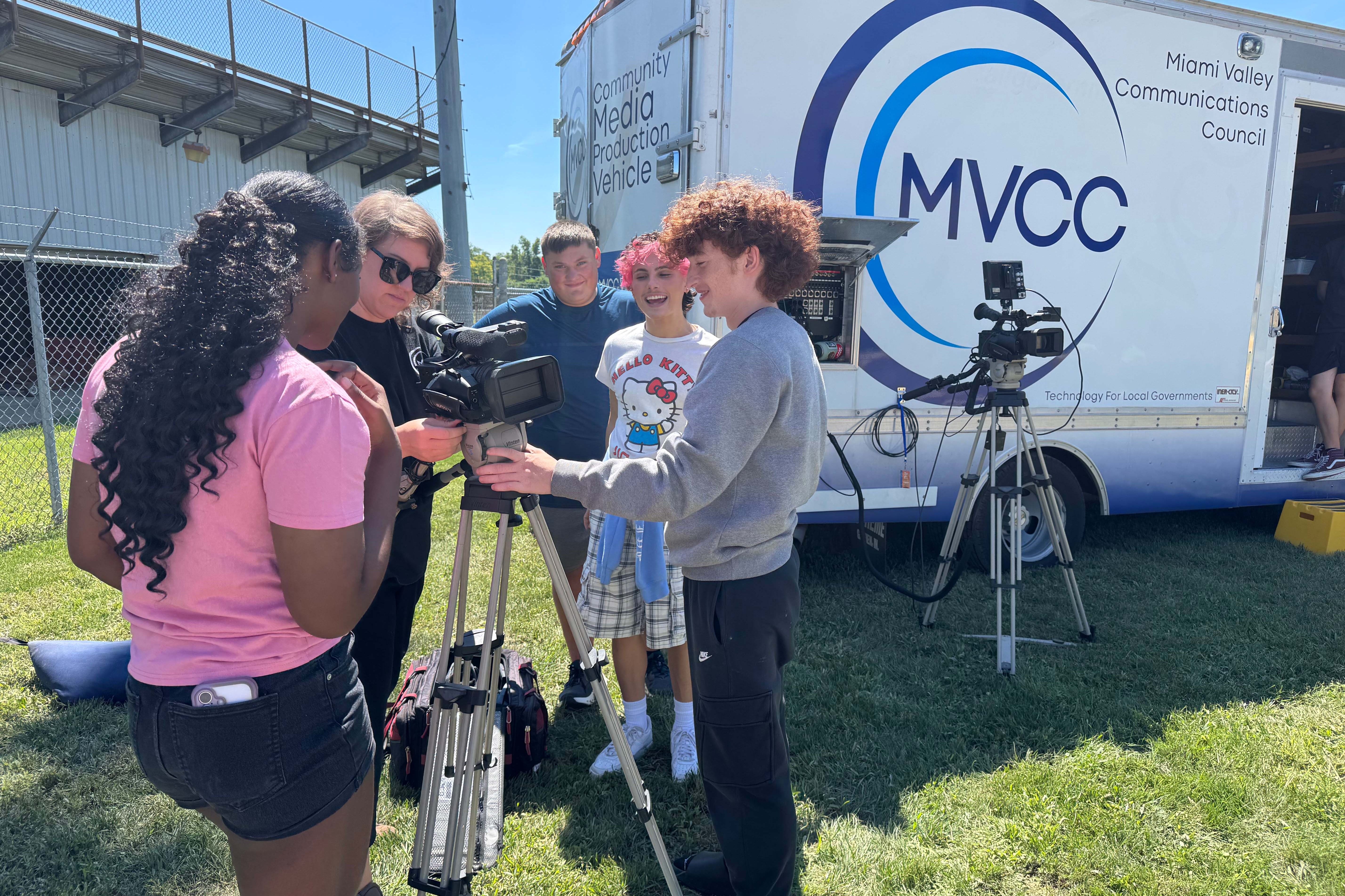 A group of people are filming with equipment on a grassy area in front of a van.