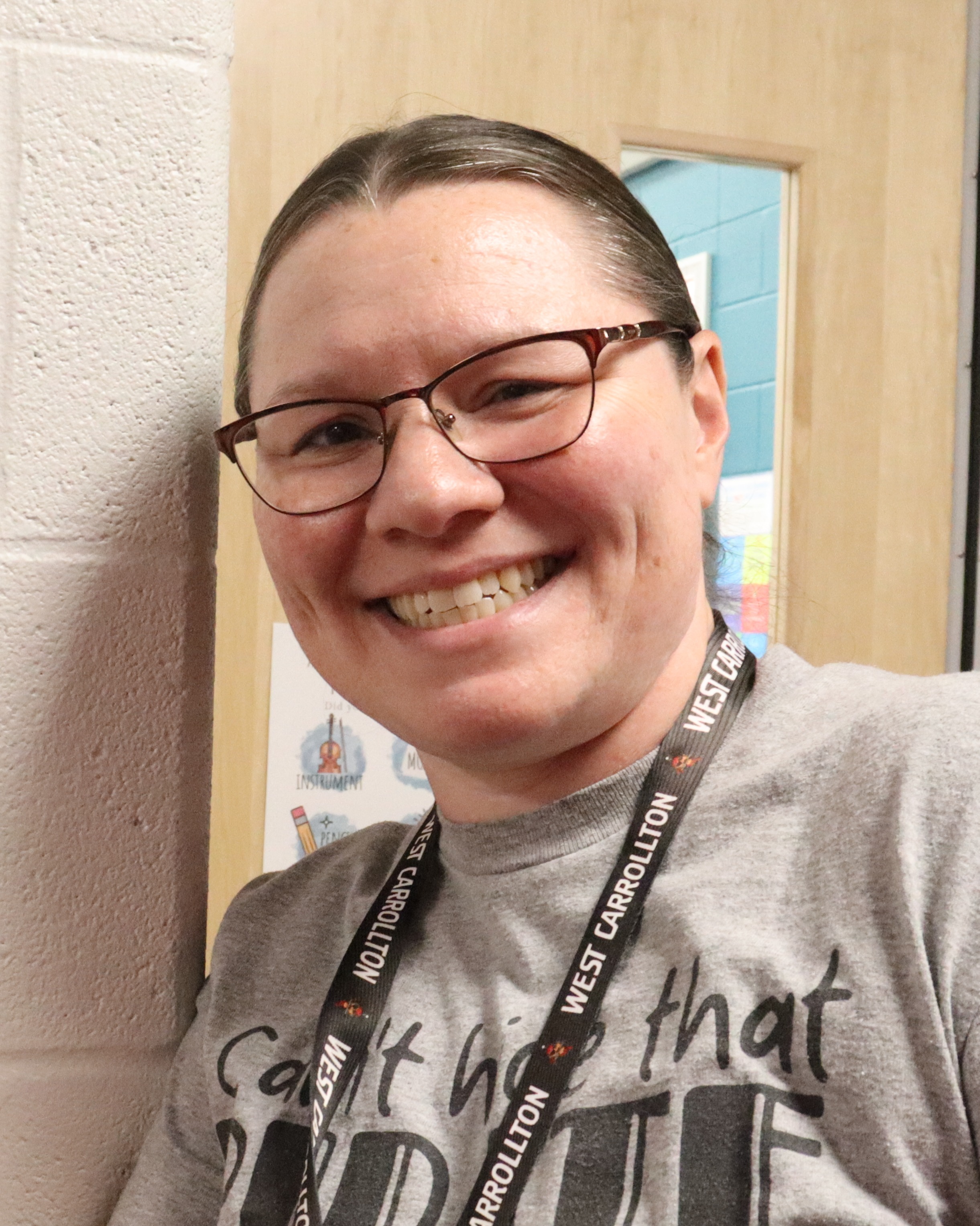 Person wearing glasses and a gray shirt stands near a white door and points to a room sign.
